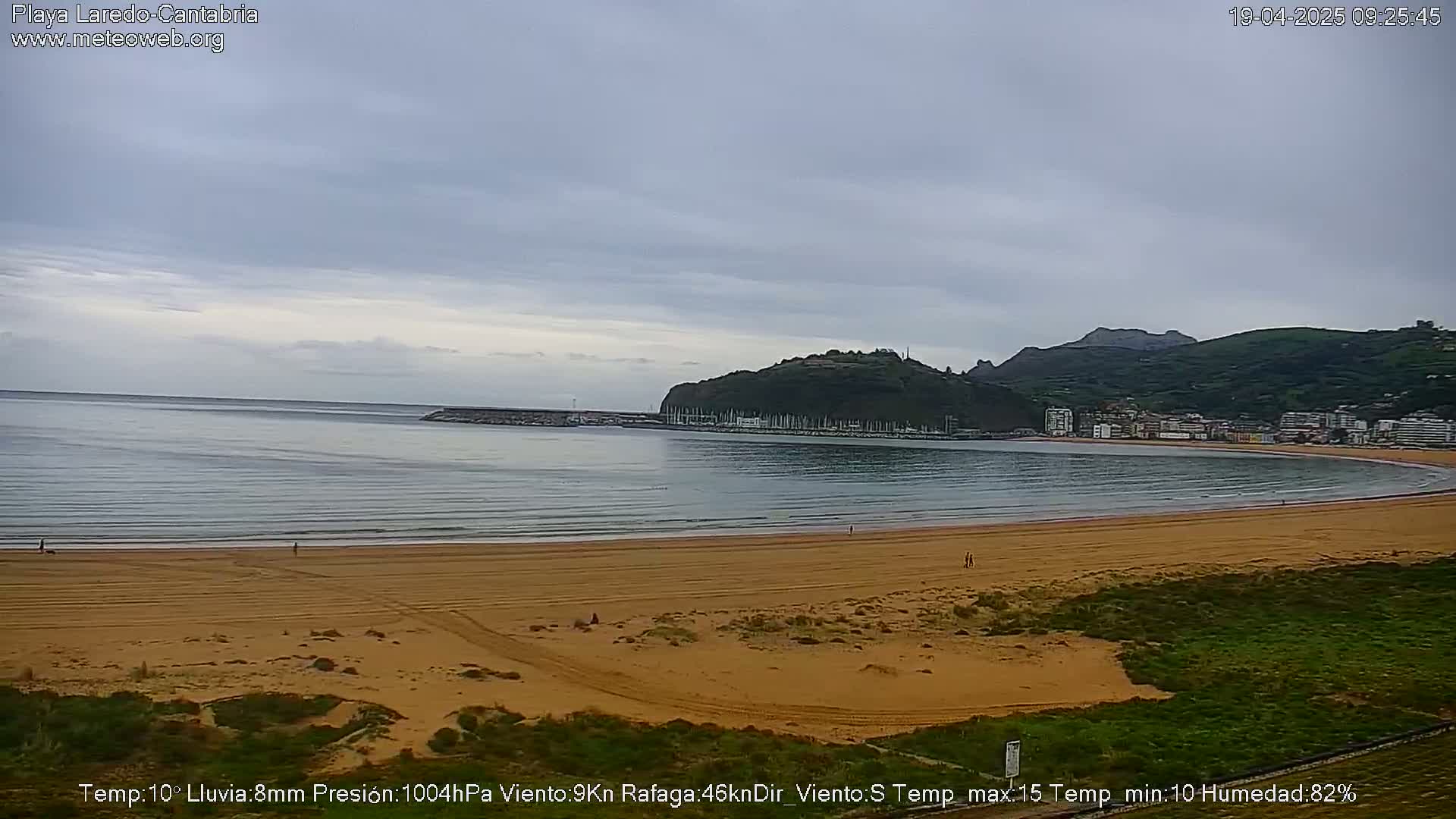 A sandy beach curves around a calm bay, under an overcast sky, with a small town nestled against hills in the background.