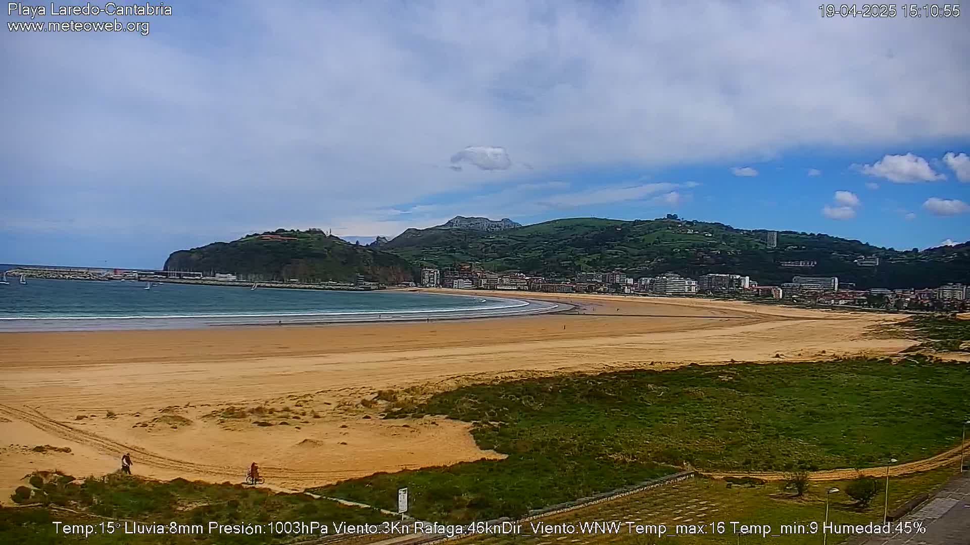 A sandy beach stretches beneath a partly cloudy sky, with a town and green hills visible in the background.
