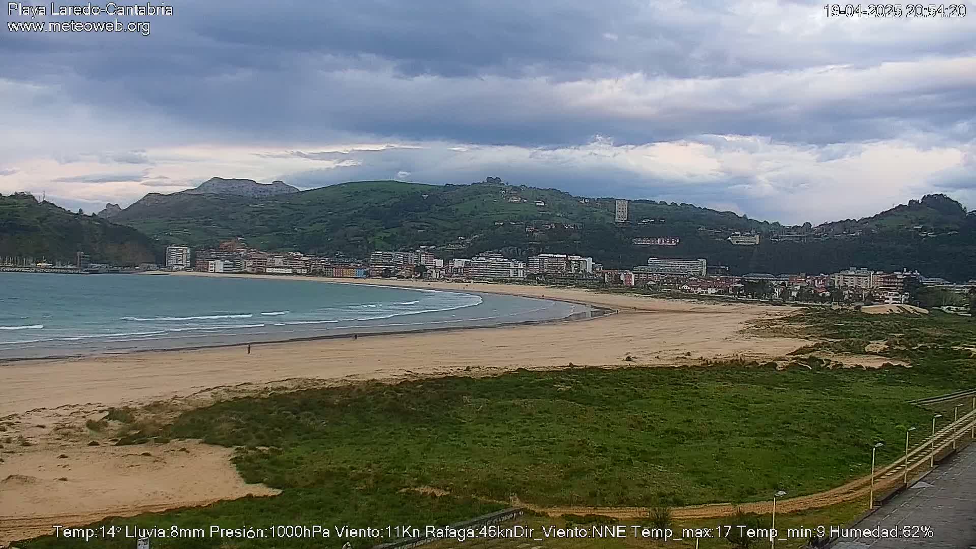 A sandy beach with gentle waves laps the shore, backed by a town nestled against a green hill under a mostly cloudy sky.