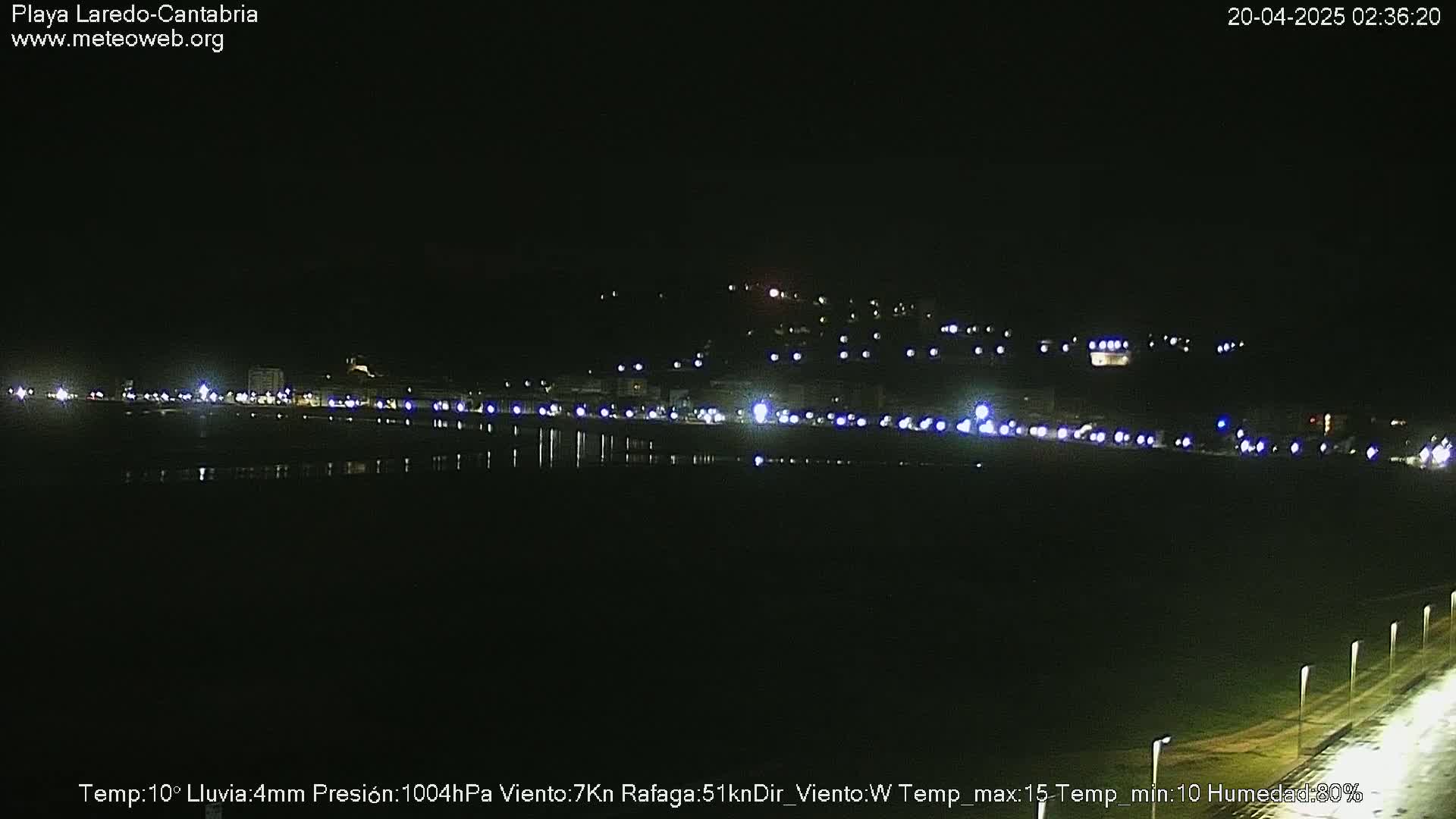 A nighttime view of a dark, calm body of water reflecting the lights of a town on a hill in the background, under a clear, dark sky.