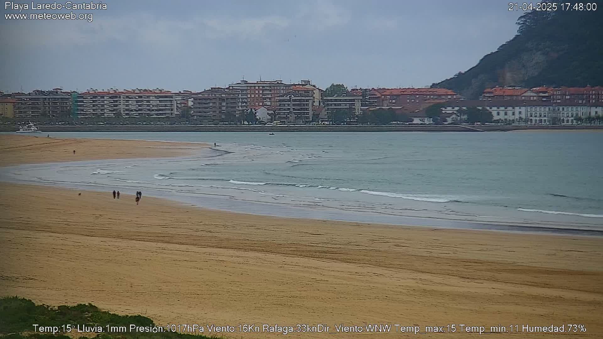 A sandy beach with gentle waves laps the shore, under an overcast sky, with buildings and a hill visible in the distance.