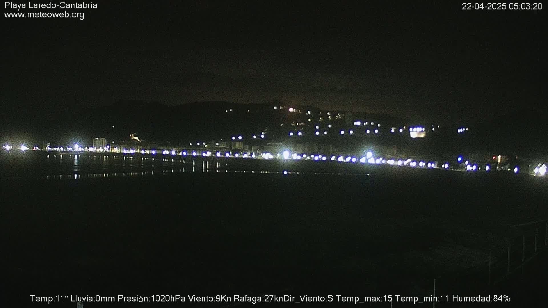 A nighttime view of a coastal town with lights reflecting on calm water under a clear, dark sky.