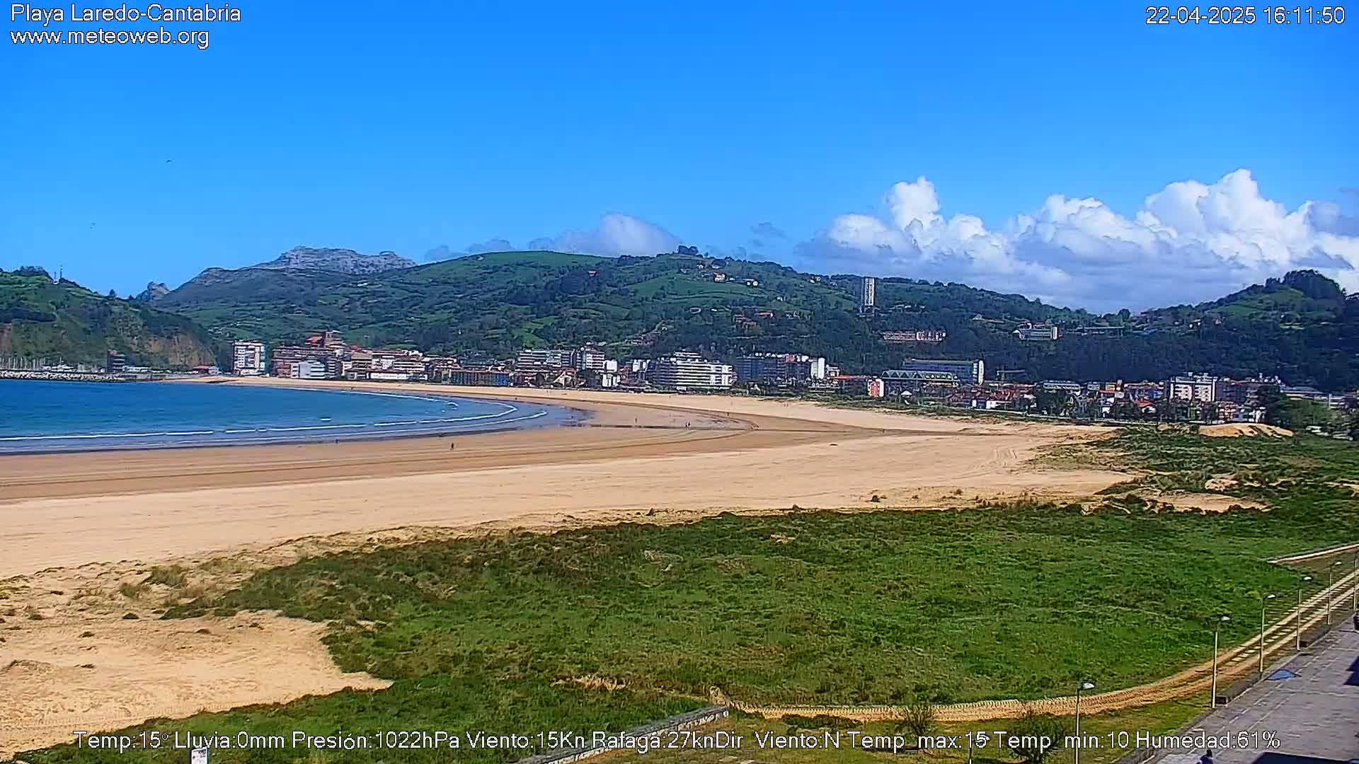 A sandy beach curves along a coastline, backed by a town nestled against verdant hills under a partly cloudy blue sky.