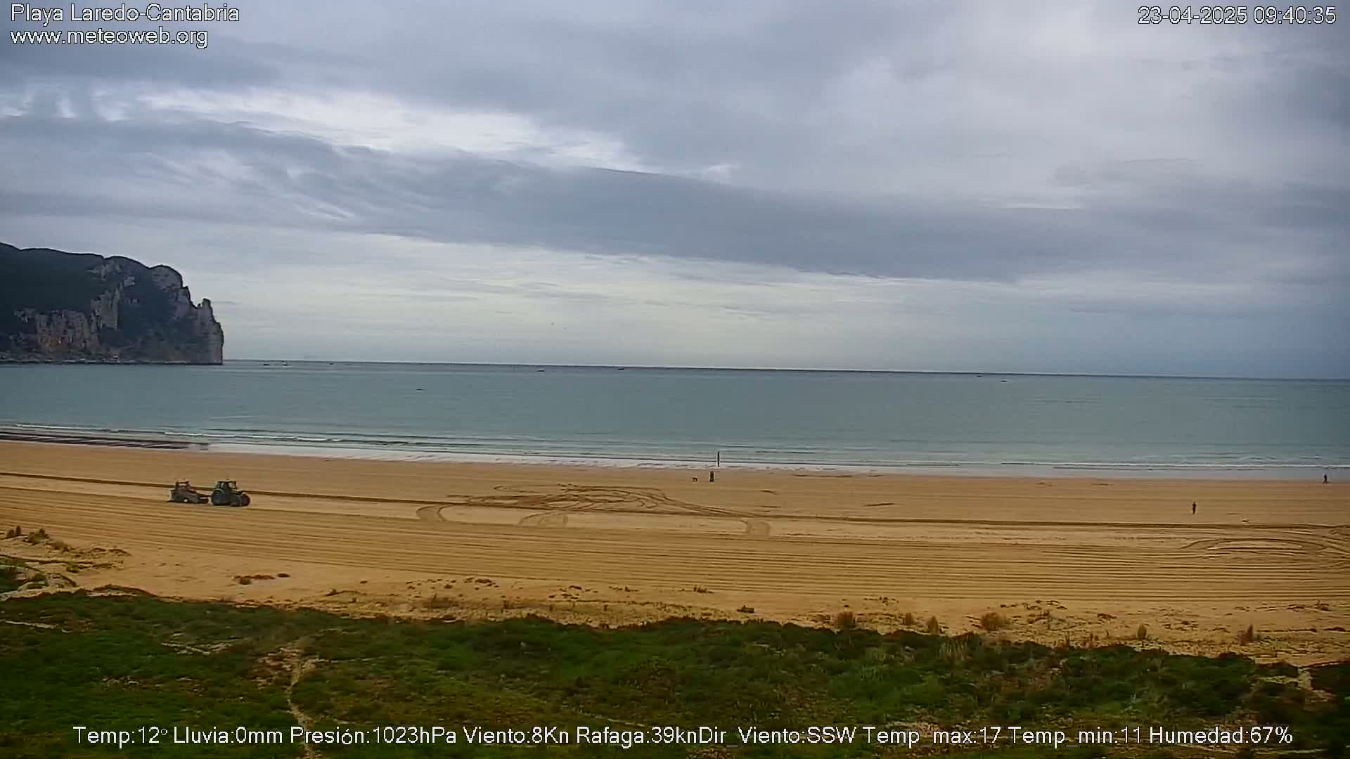 A sandy beach with tire tracks and a tractor is visible, under an overcast sky, with calm waves lapping the shore and a large cliff in the distance.