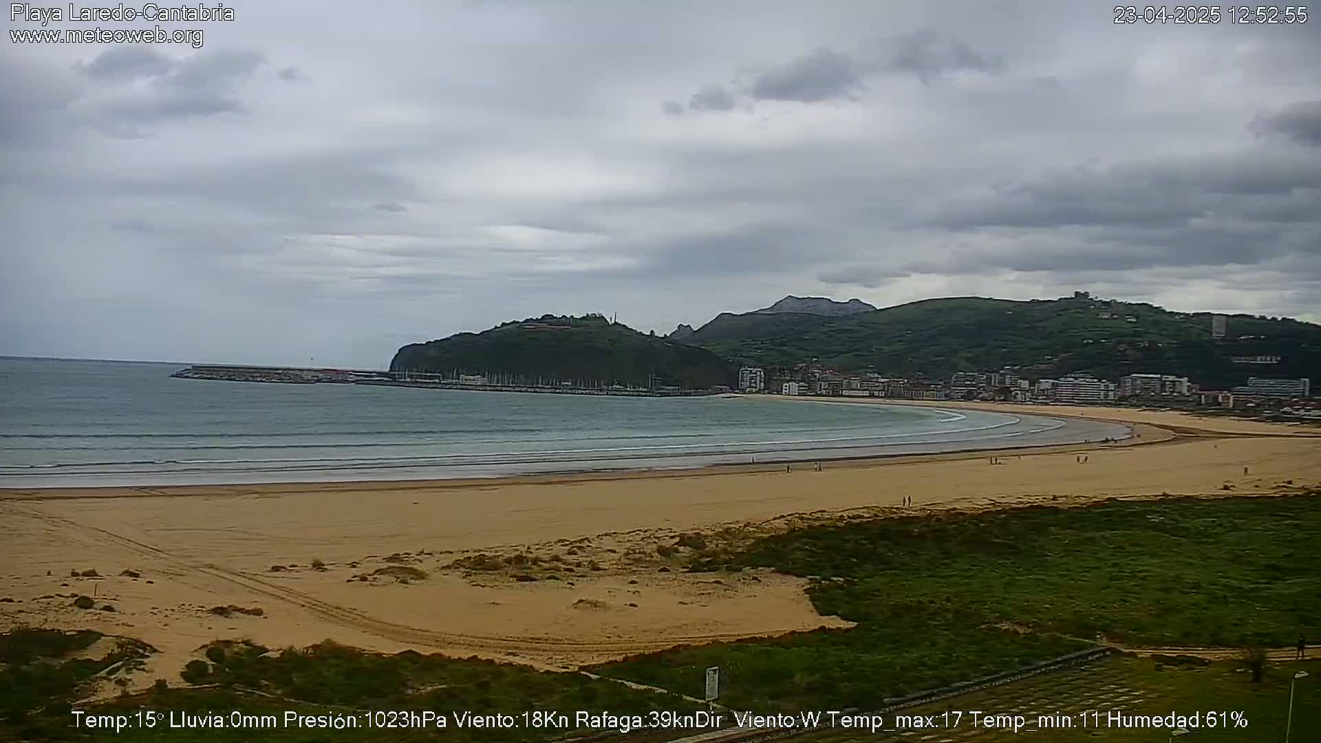 A sandy beach curves along a calm ocean under an overcast sky, backed by a town nestled against green hills.