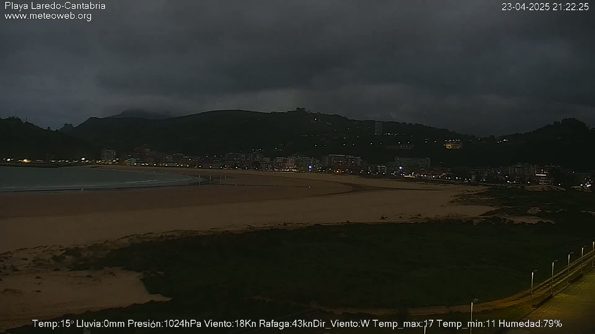 A dark, overcast night scene shows a beach at low tide with a town and hills in the background.