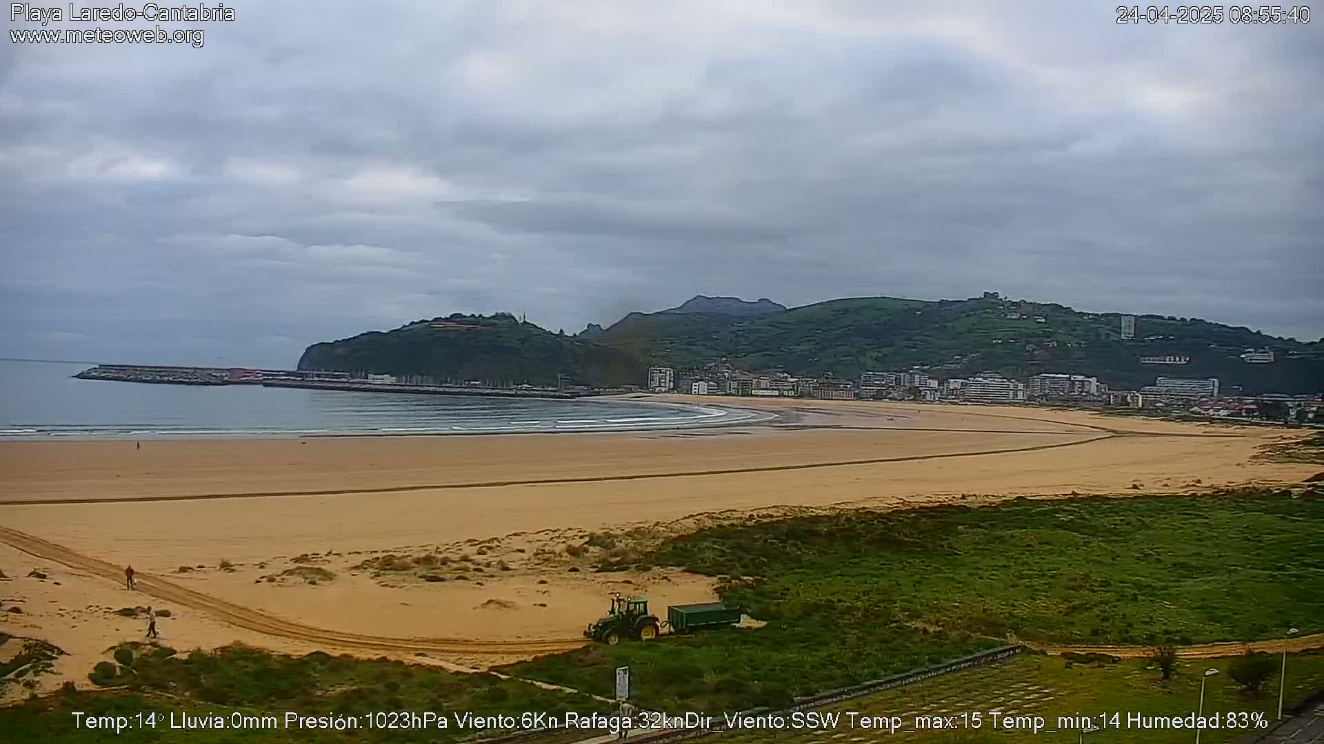 A sandy beach stretches along a calm ocean under an overcast sky, with a town nestled against hills in the background, and a tractor visible near the lower edge of the beach.