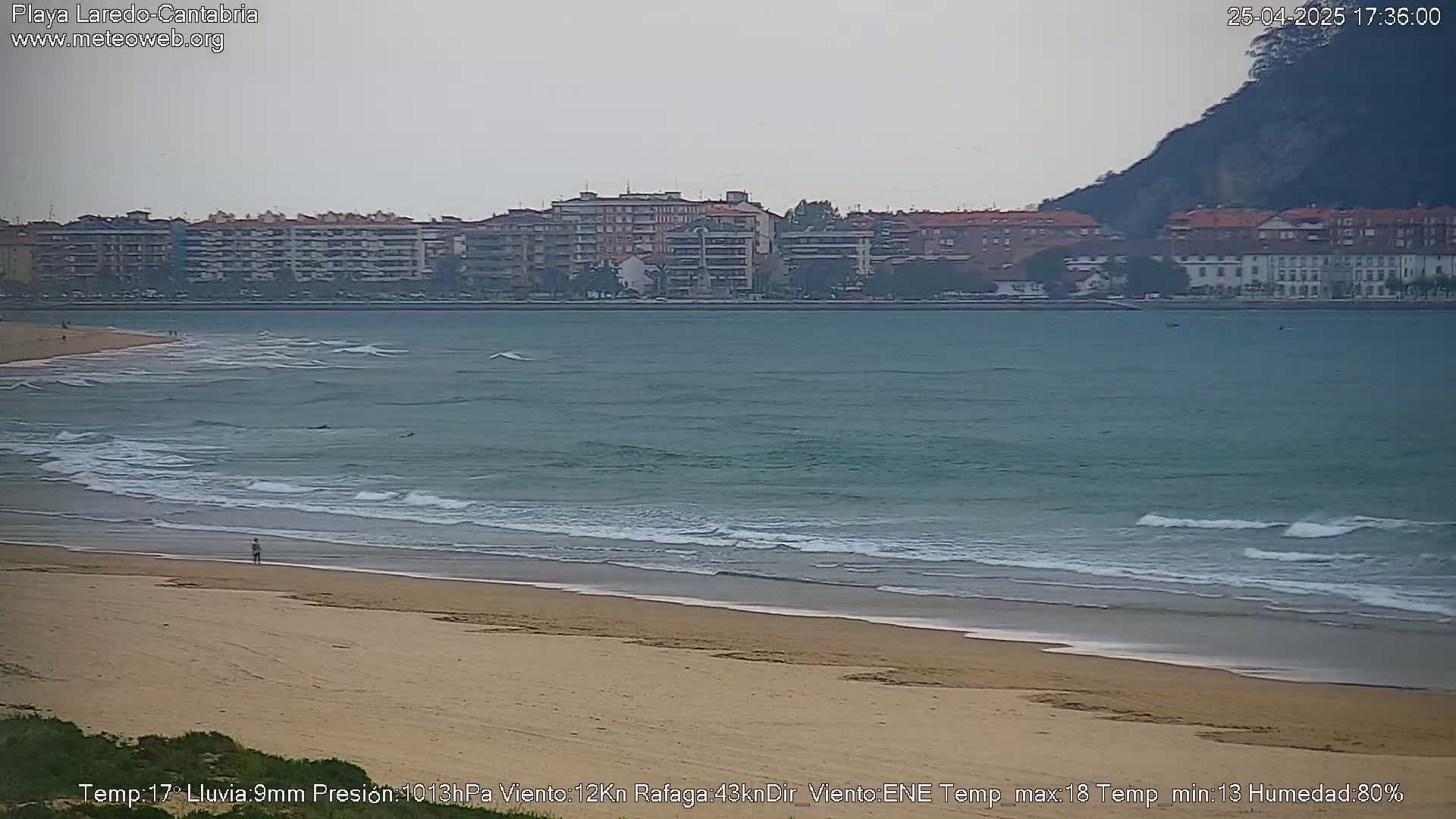 A sandy beach with gentle waves laps against the shore, under an overcast sky, in front of a town with multi-story buildings and a hill in the distance.
