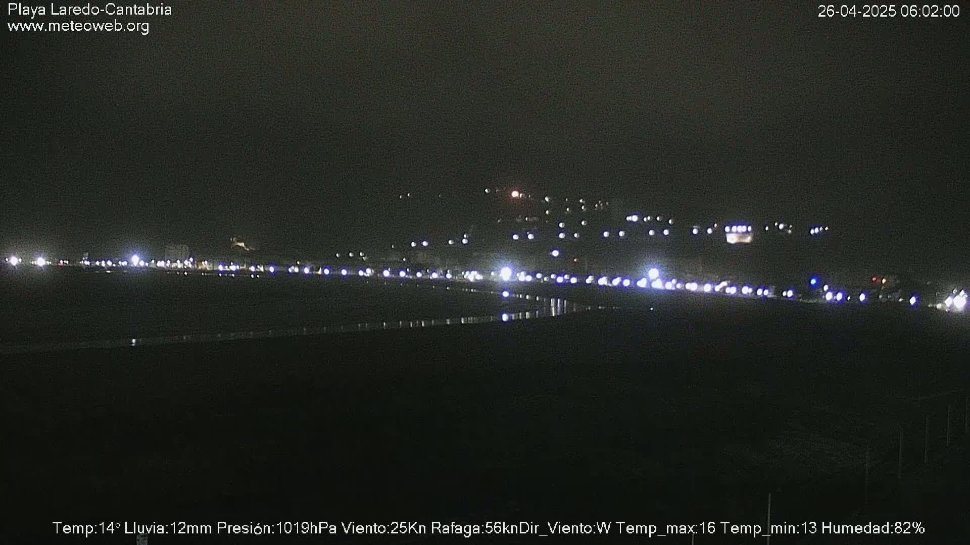 A nighttime view of a coastal town with numerous lights visible, under rainy conditions.