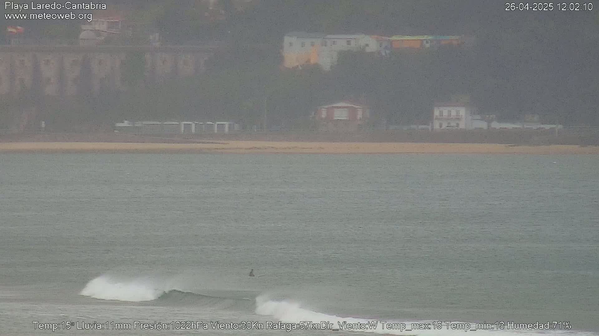 A person surfs in the ocean near a beach with buildings and hills in the background on an overcast day with light rain.