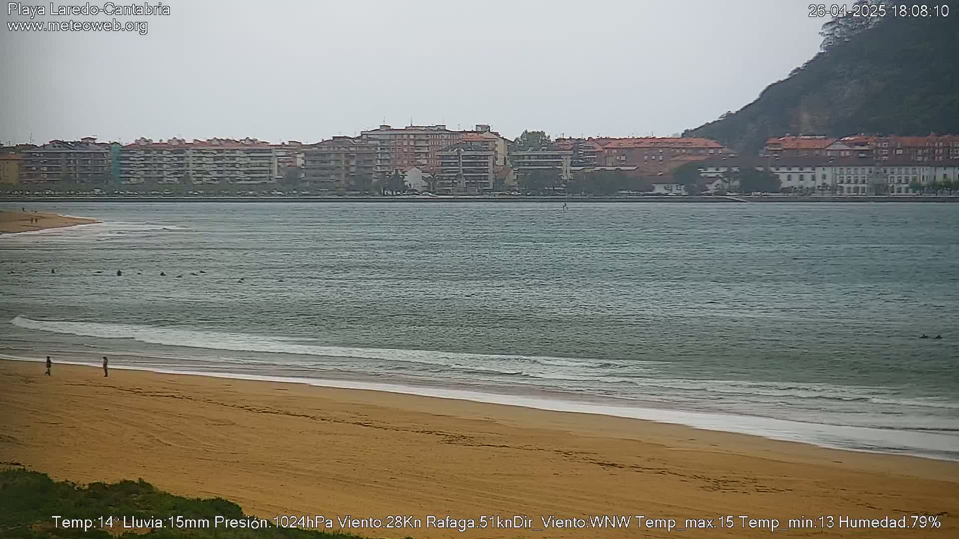 A mostly cloudy day shows a sandy beach with a few people on it, gentle waves lapping the shore, and a calm bay with buildings and a hill in the background.