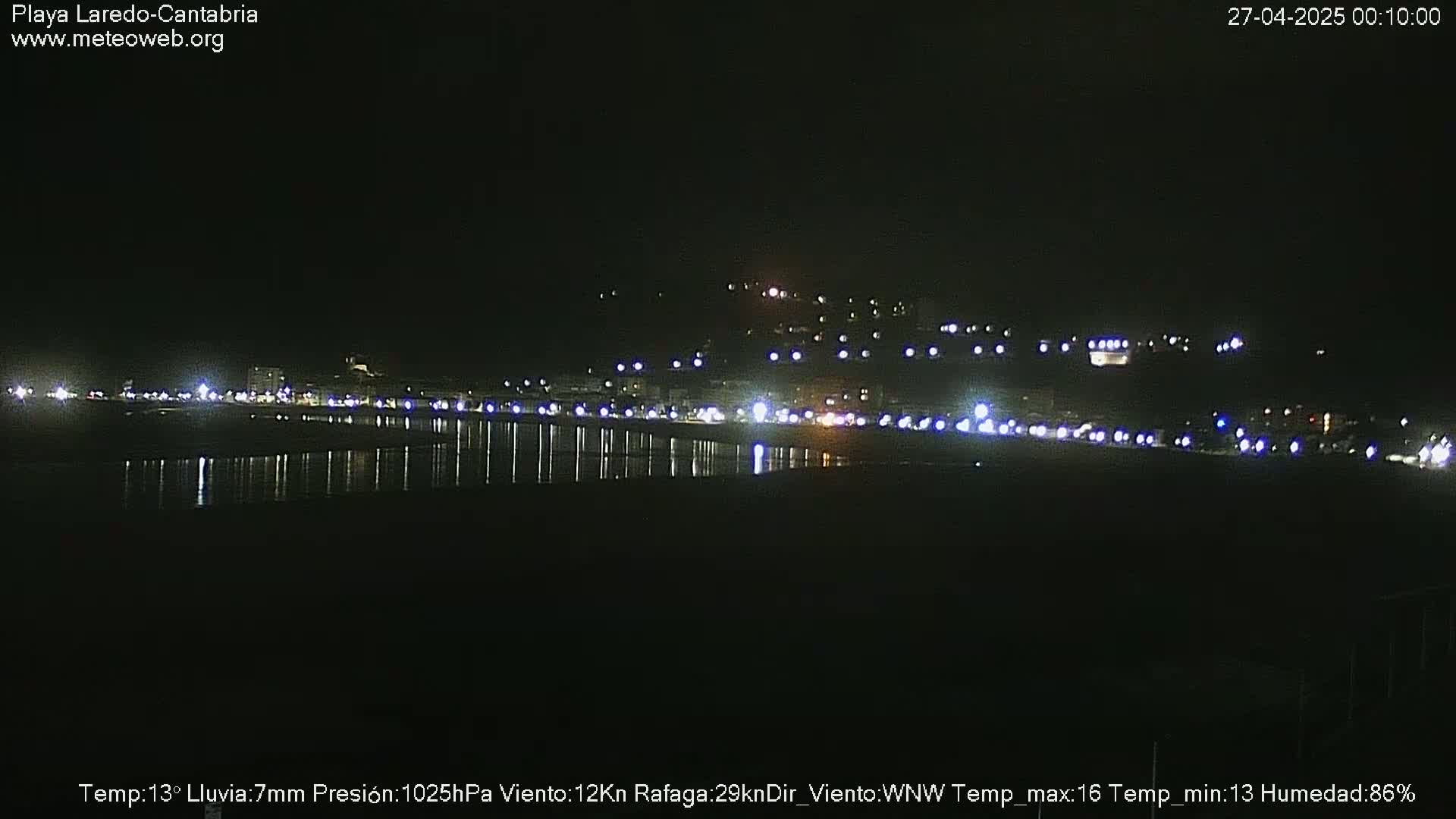 A nighttime view of a town's lights reflected in dark water under a dark sky, with light rain.