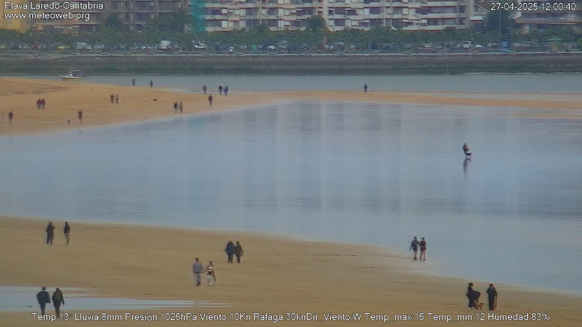 A sandy beach with scattered people stretches along a calm, light-blue body of water under a cloudy sky.