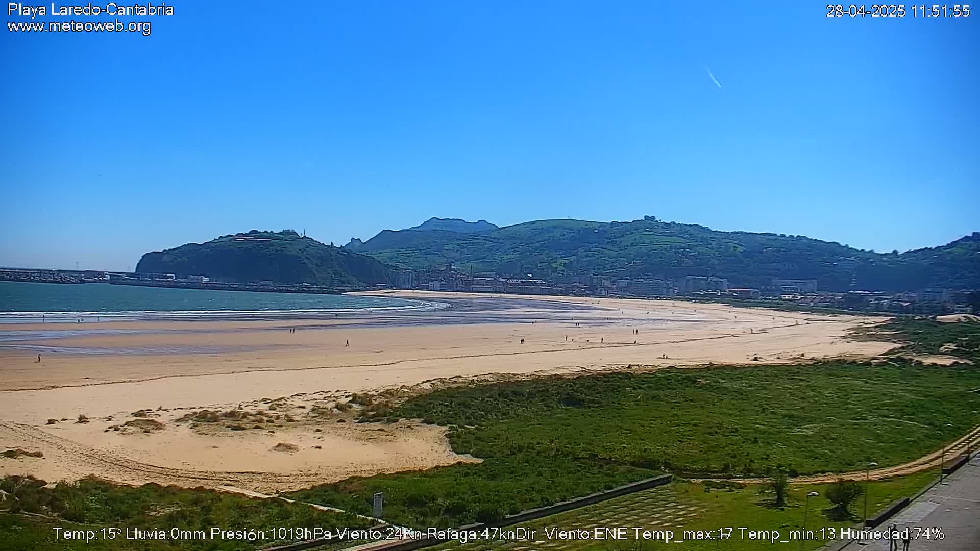 A sandy beach stretches along a calm ocean under a clear blue sky, backed by green hills and a town.