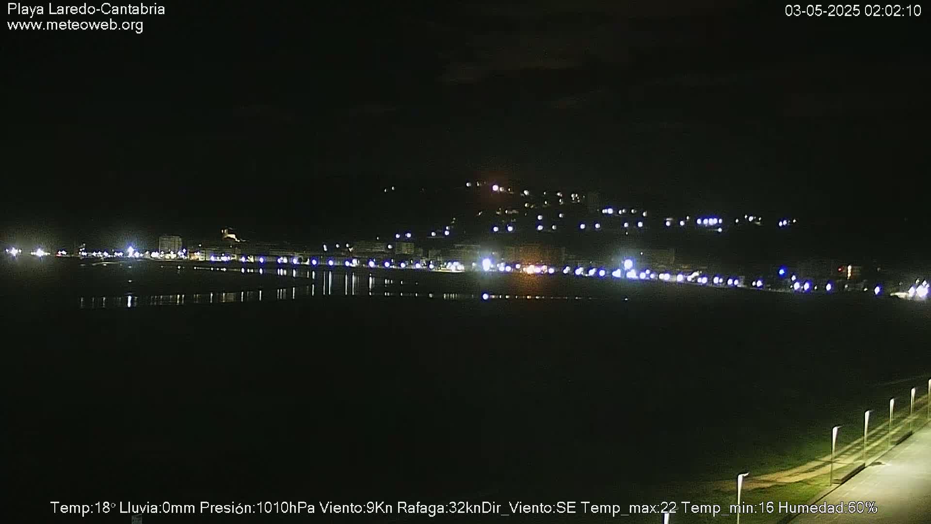 A nighttime view of a coastal town with its lights reflecting on calm water under a clear sky.