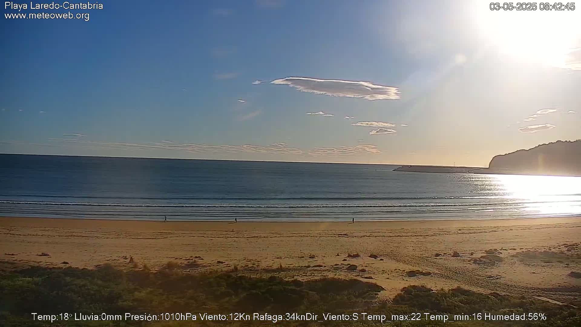 A sandy beach with gentle waves under a mostly clear sky, with some scattered clouds.