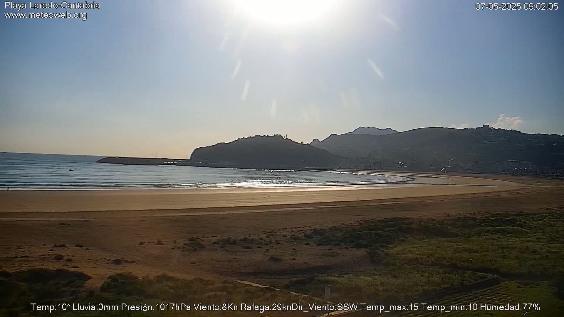 A sandy beach curves around a calm bay under a bright, sunny sky, backed by hills.