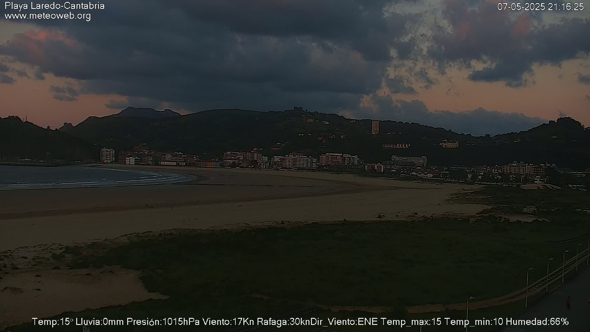 A mostly cloudy evening sky over a sandy beach and town nestled against dark hills.