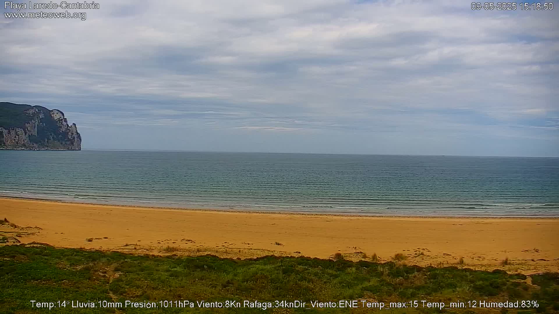 A sandy beach meets a calm ocean under a mostly cloudy sky, with a large cliff visible in the distance.