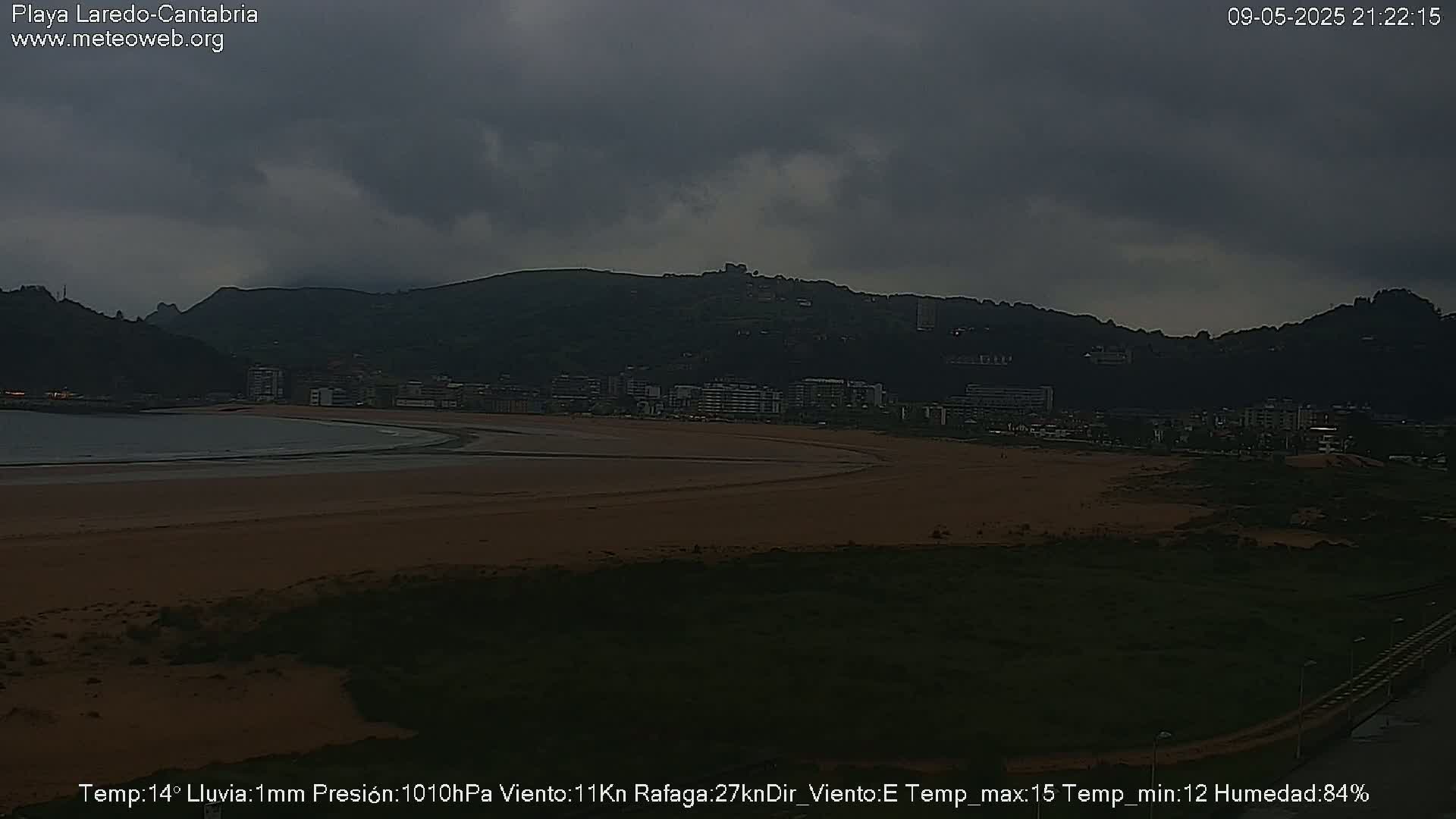 A dark, overcast sky hangs over a sandy beach and town backed by hills.
