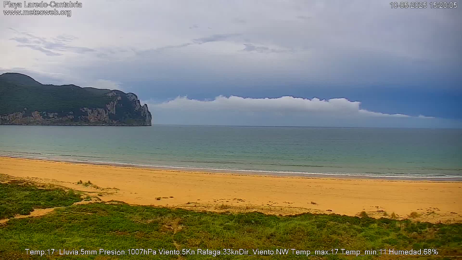 A sandy beach stretches to a calm ocean under a partly cloudy sky, with a large, dark cliff visible in the distance.