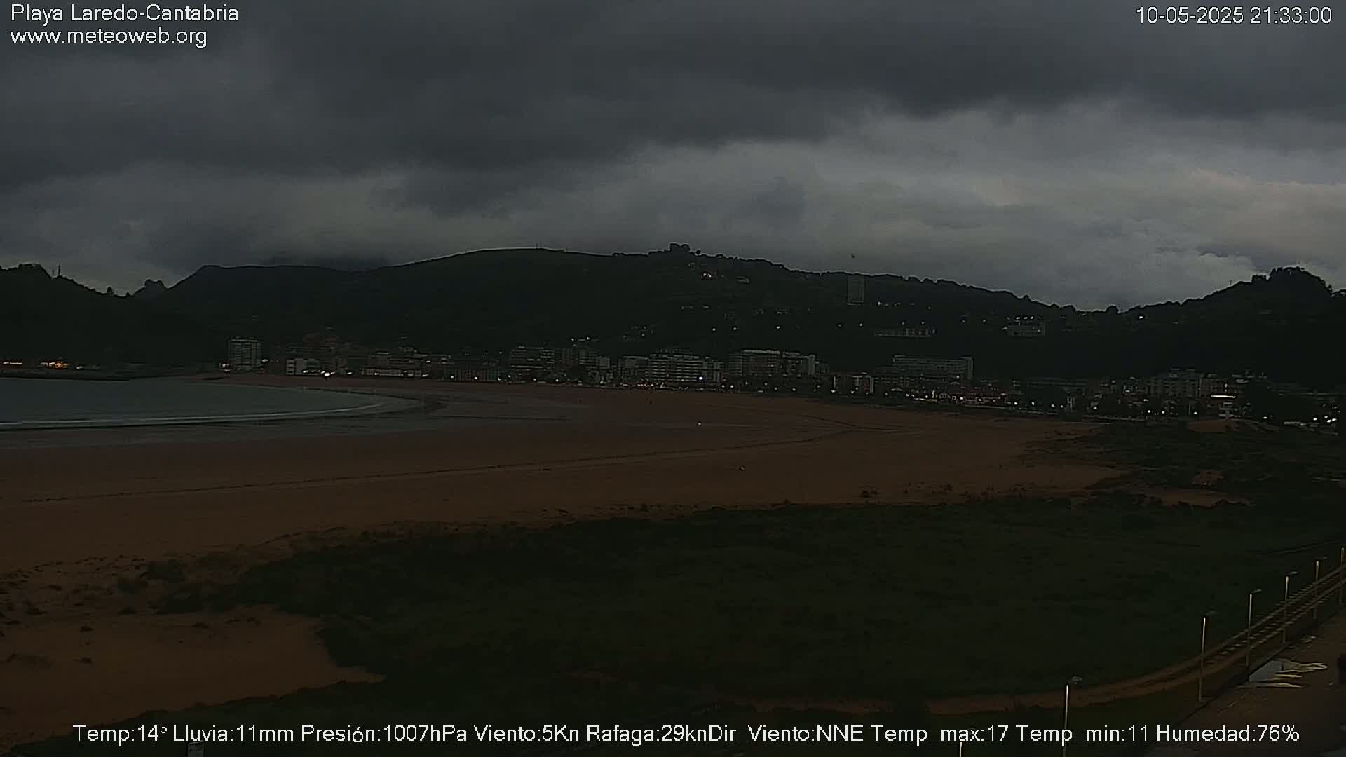 A dark, overcast sky hangs over a beach and town nestled against a mountain range under rainy conditions.