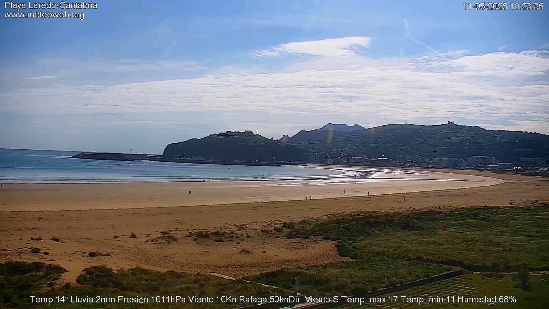 A sandy beach curves around a calm bay, backed by green hills under a partly cloudy sky.