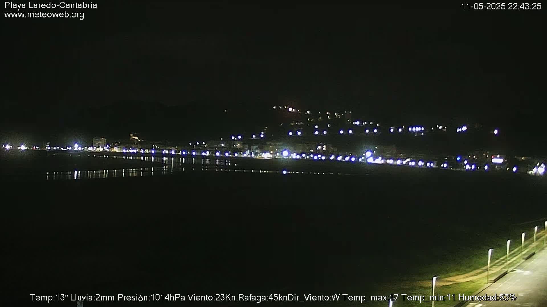 A nighttime coastal view shows a town illuminated with lights along a dark body of water under a clear sky.