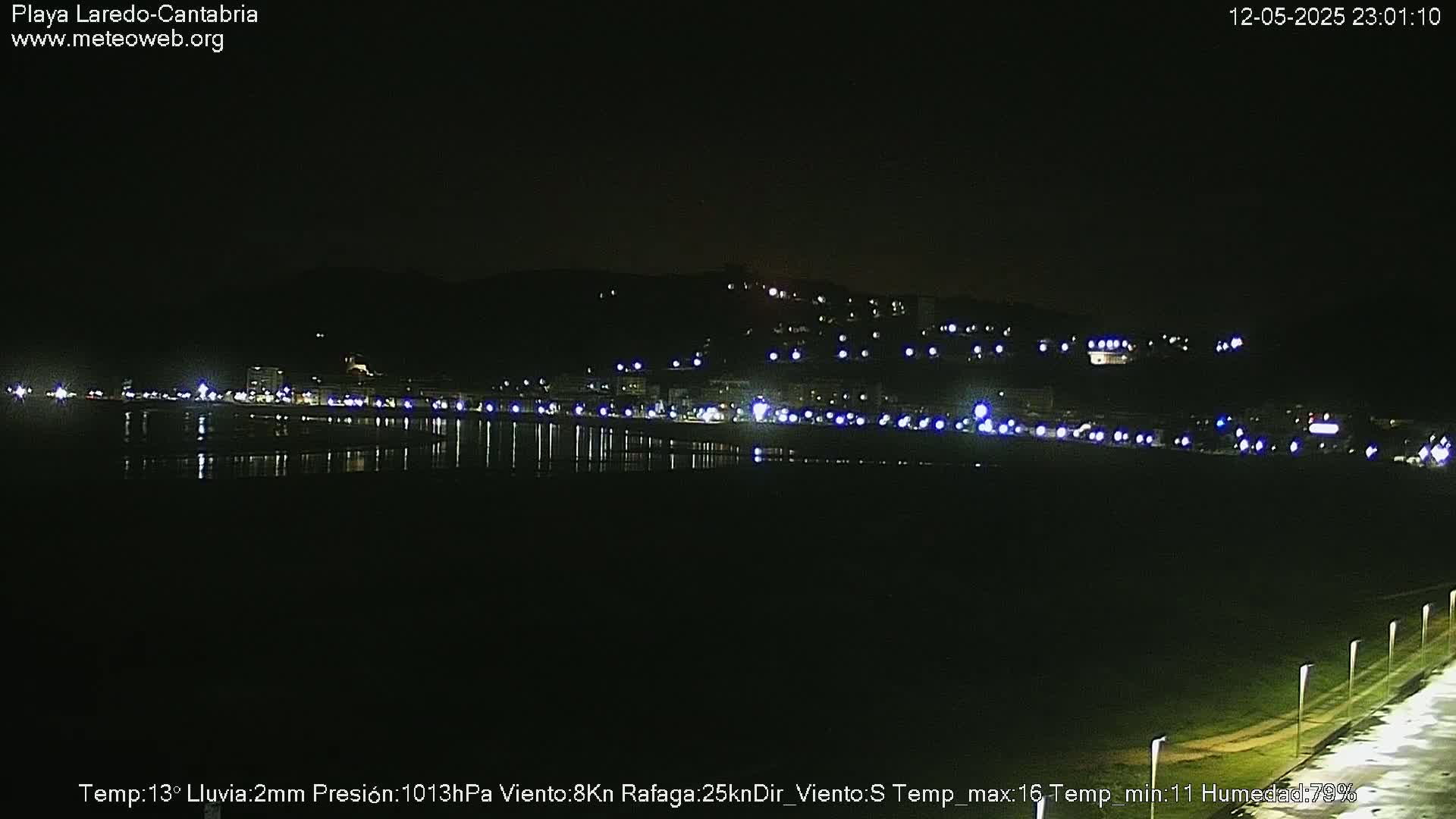 A nighttime view of a coastal town with lights reflecting on calm water under a clear, dark sky.