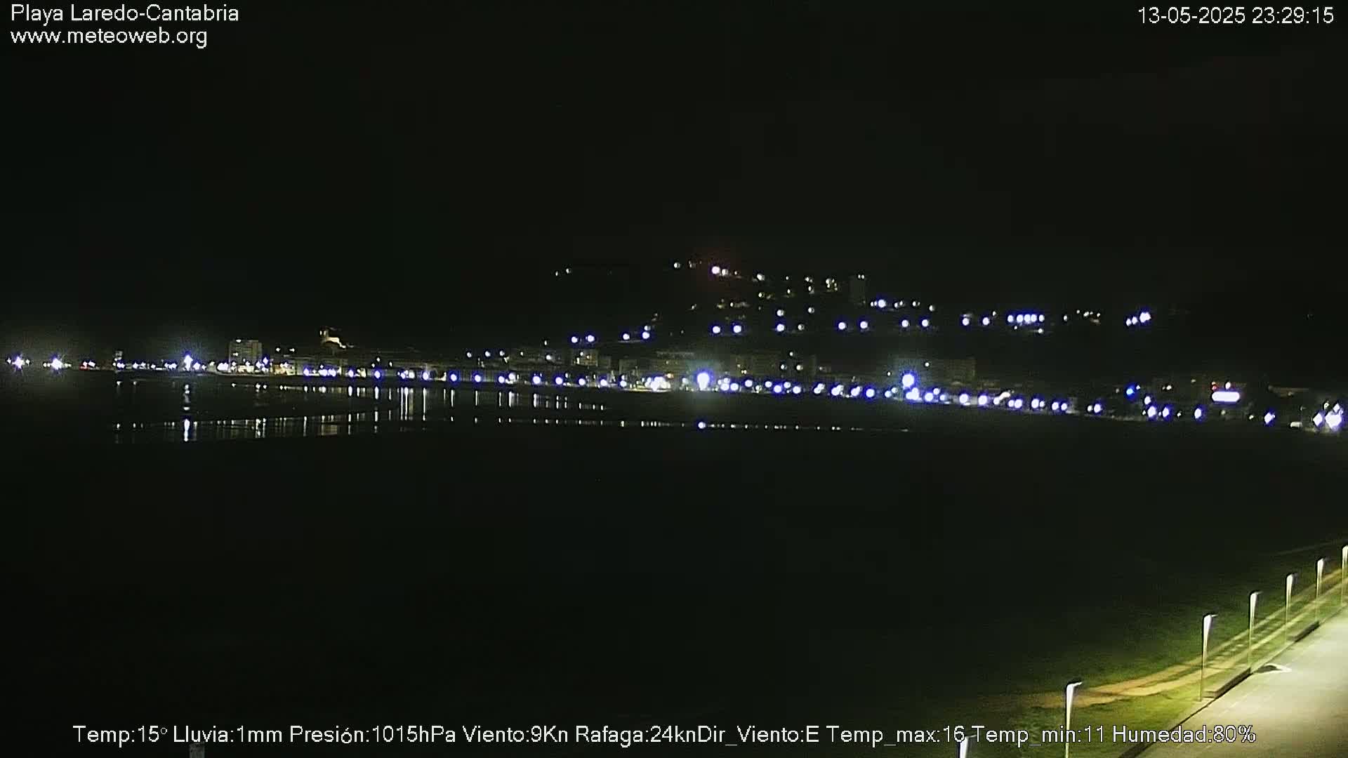 A nighttime view of a coastal town with lights reflecting on the water under a dark, rainy sky.