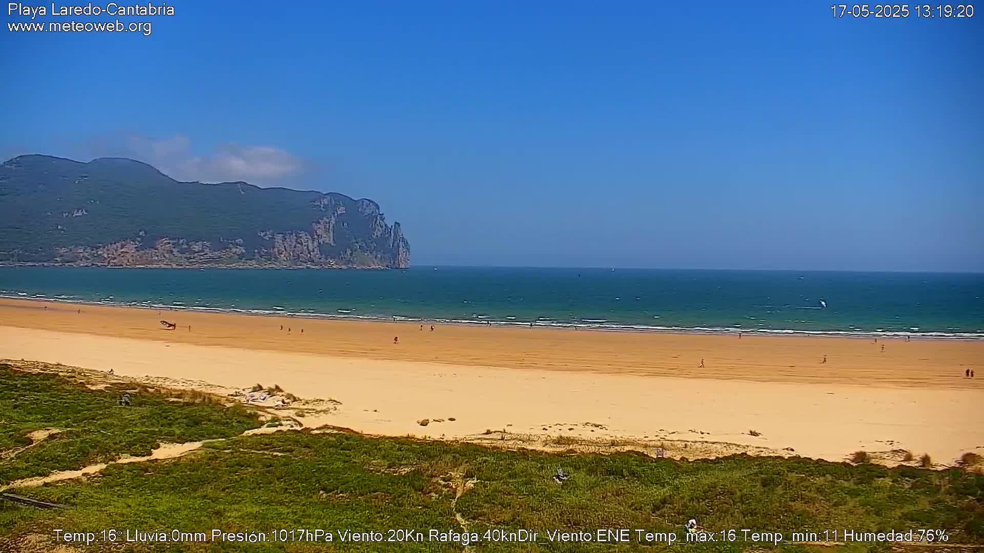 A sandy beach stretches out to a calm ocean under a clear blue sky, with a mountainous backdrop.
