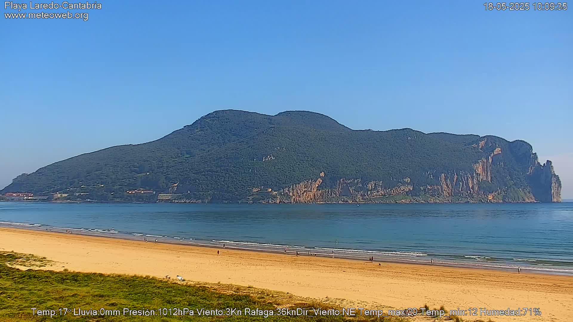 A sandy beach stretches toward a large, verdant mountain across a calm ocean under a clear blue sky.