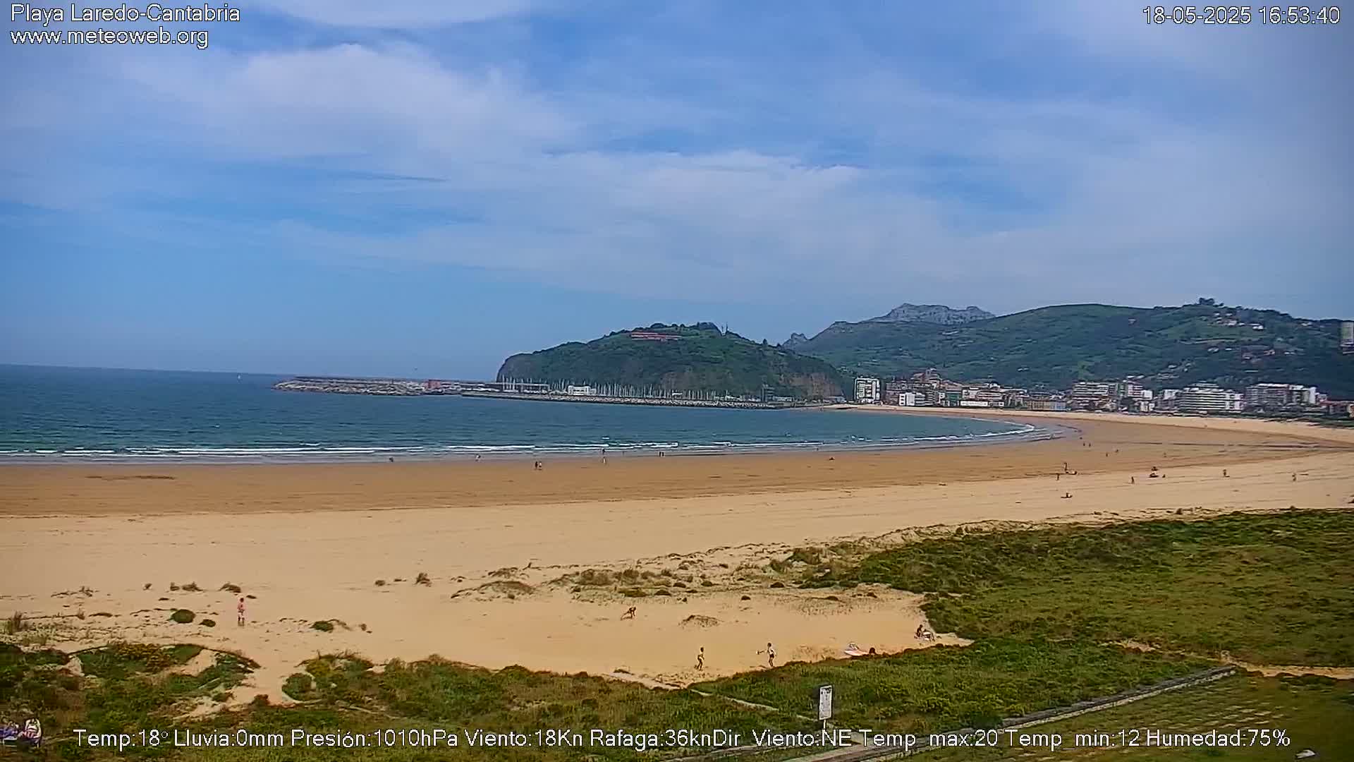 A long sandy beach stretches out to a calm ocean under a mostly clear sky, with a town and green hills in the background.