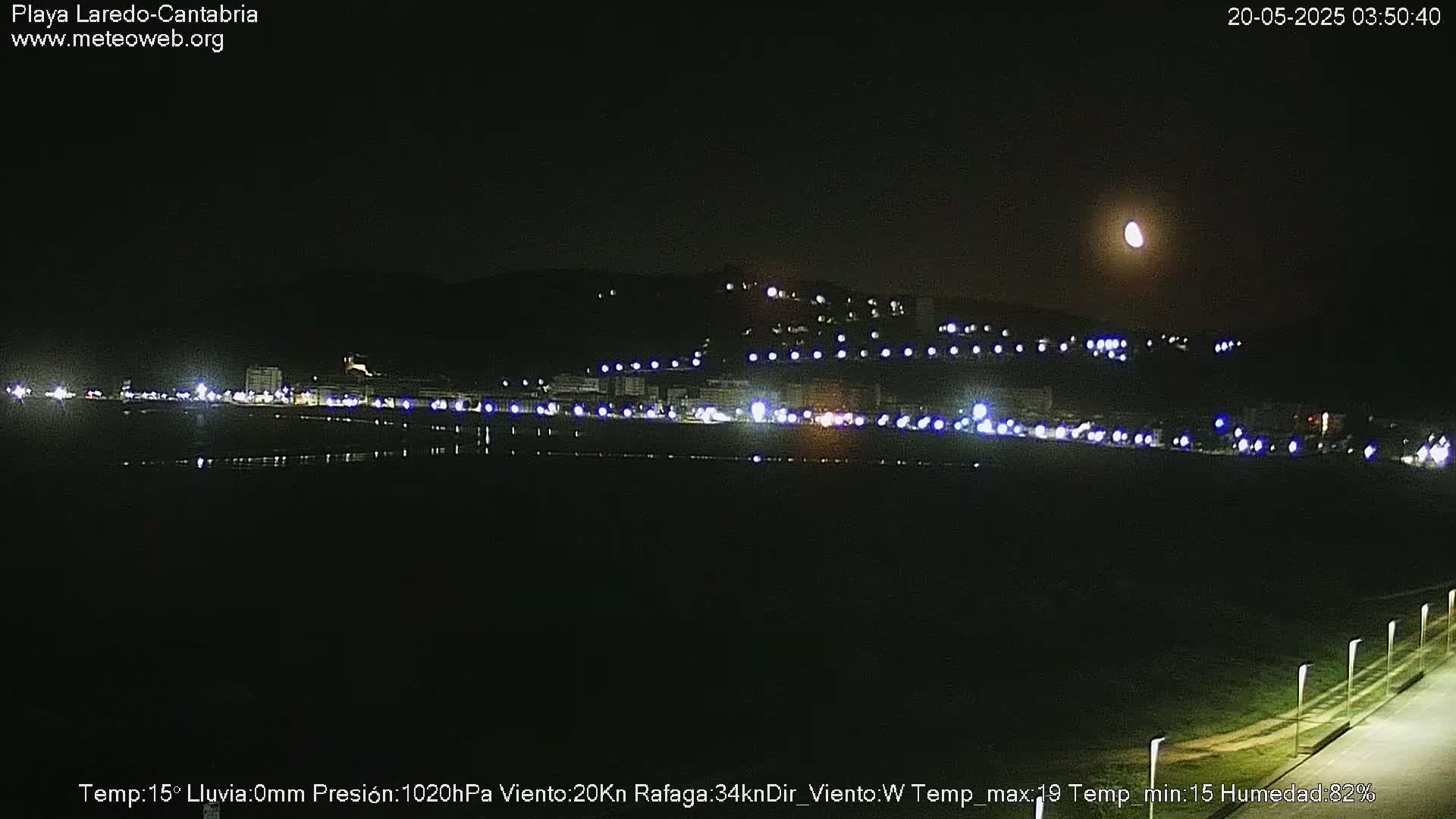 A nighttime view of a city's coastline illuminated by lights, under a clear sky with a visible moon.