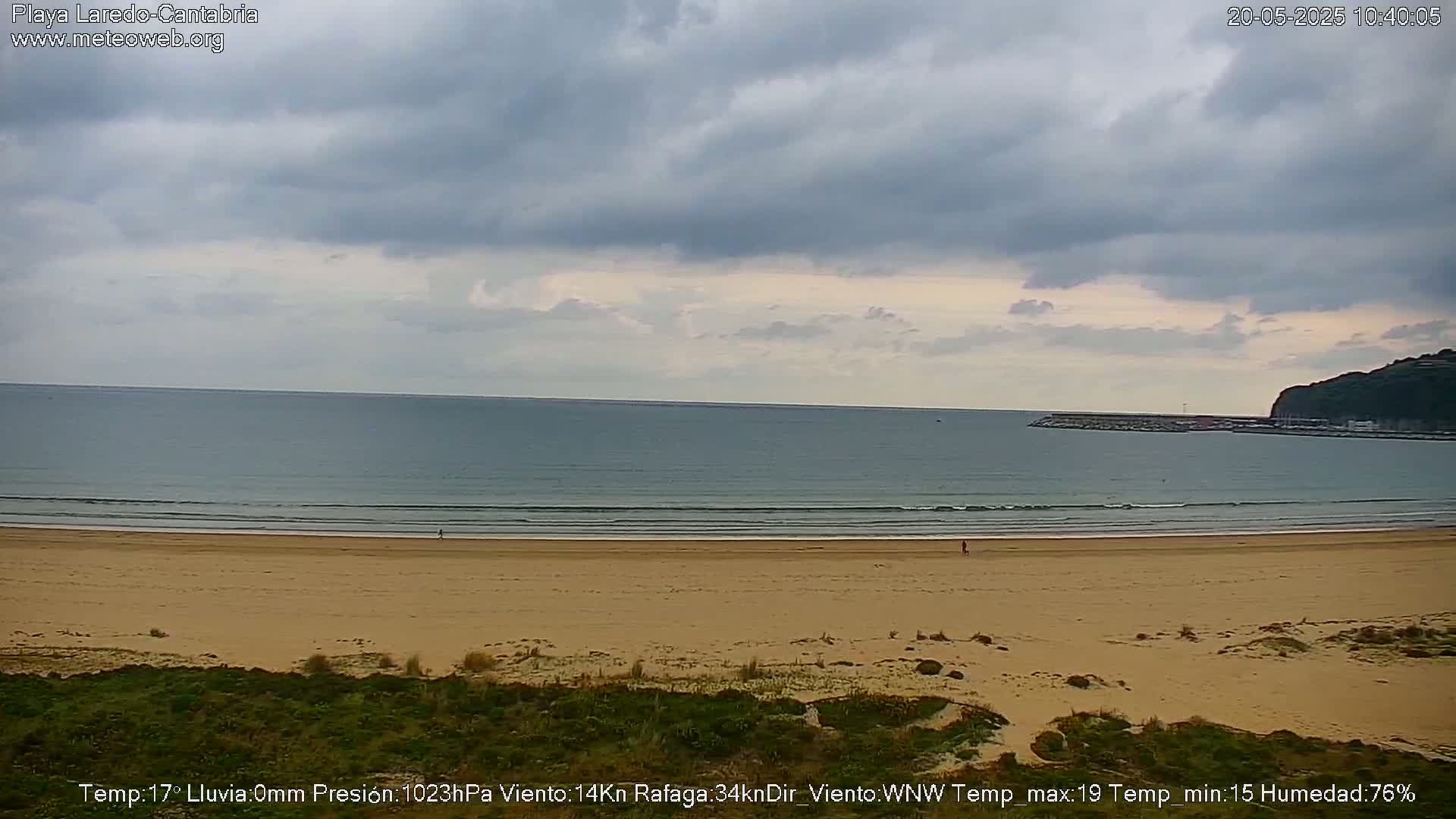 A mostly cloudy sky hangs over a wide sandy beach and calm ocean, with a distant headland visible.