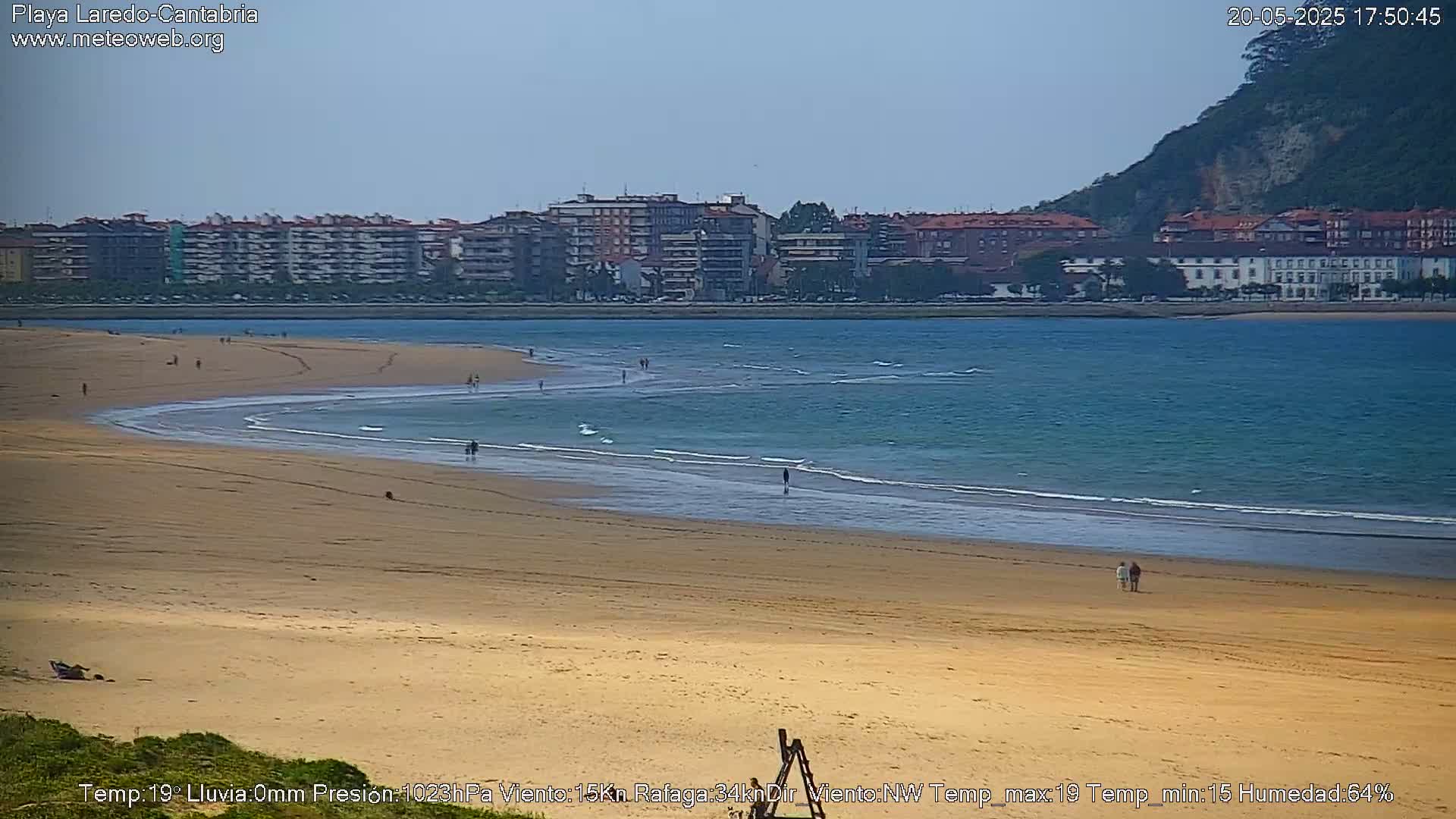 A sandy beach with gentle waves laps at the shore, under a mostly clear sky, with buildings and a hill visible in the distance.