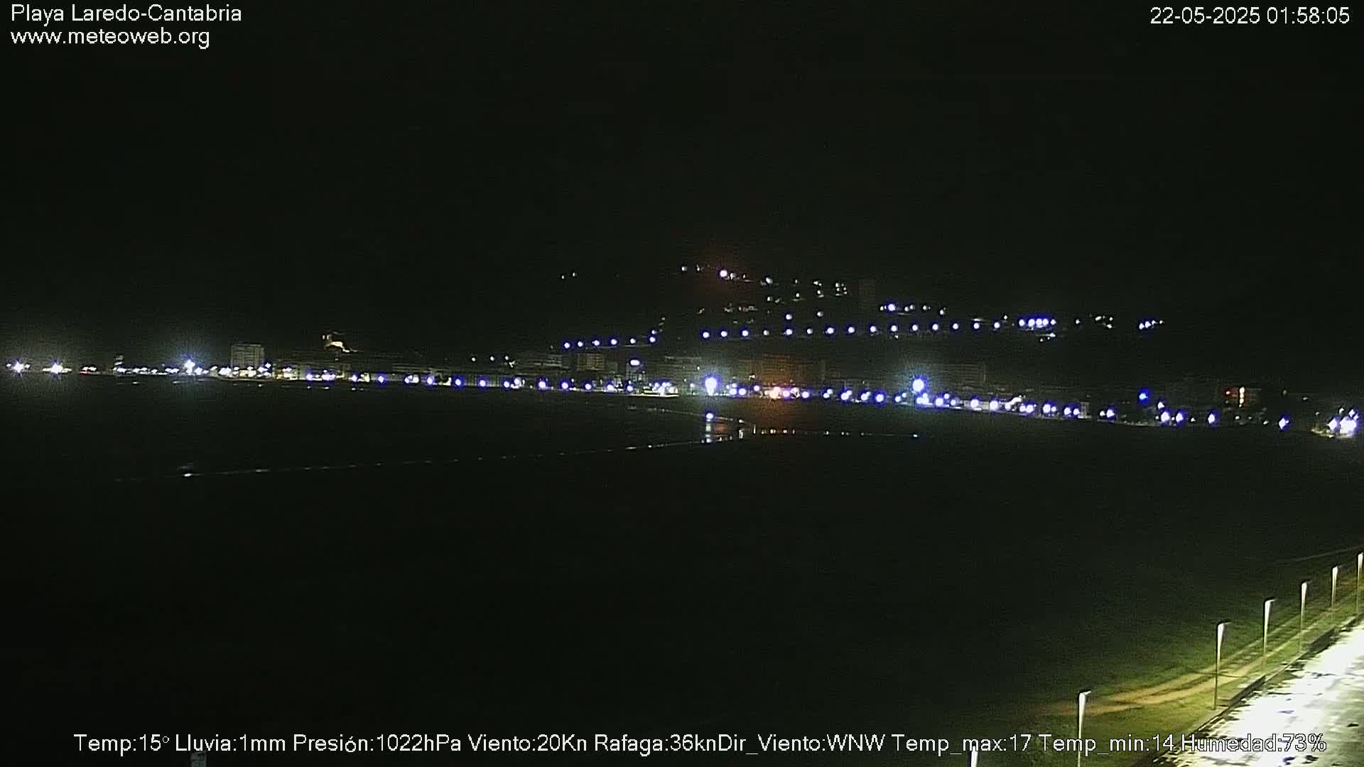 A nighttime view of a coastal town with streetlights illuminating a dark beach and hillside under rainy conditions.