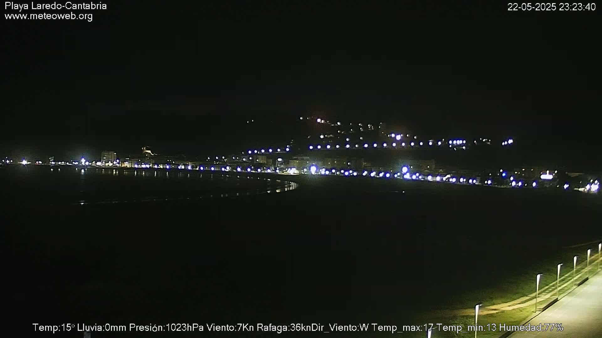 A nighttime long shot of a coastal town illuminated by streetlights, with calm waters and a dark sky.