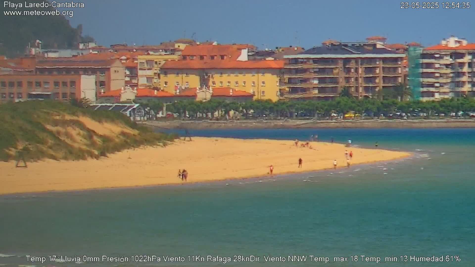 A sandy beach with a few people on it stretches into a calm body of water in front of a town of multi-story buildings under a clear sky.