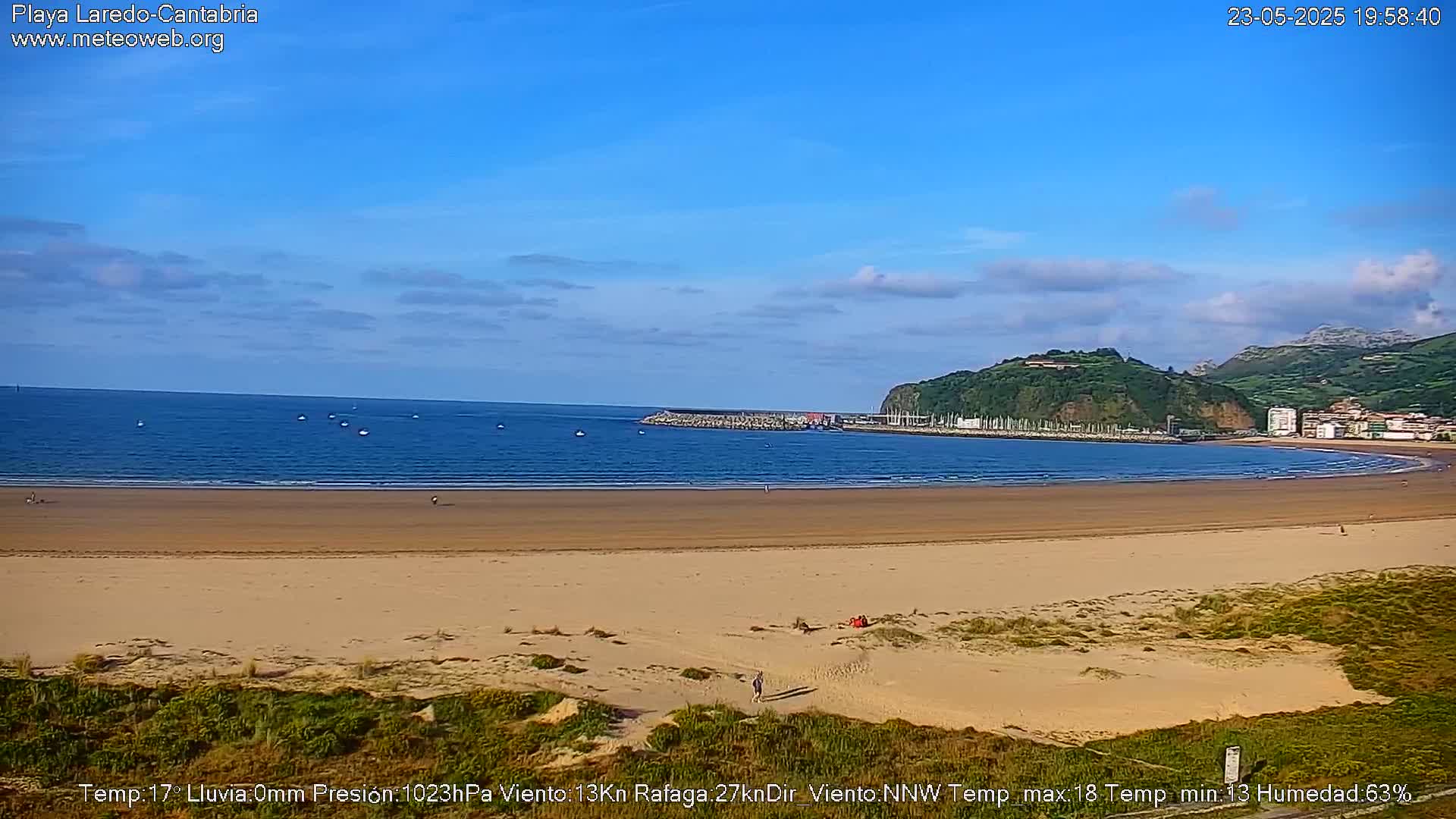 A wide sandy beach curves gently around a calm bay under a mostly clear blue sky, with a small town visible in the background.