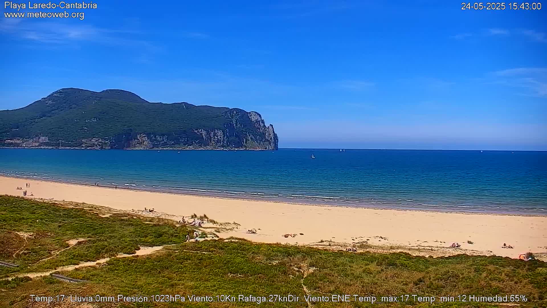 A sandy beach stretches along a calm ocean under a clear blue sky, with a large, verdant hill rising in the background.