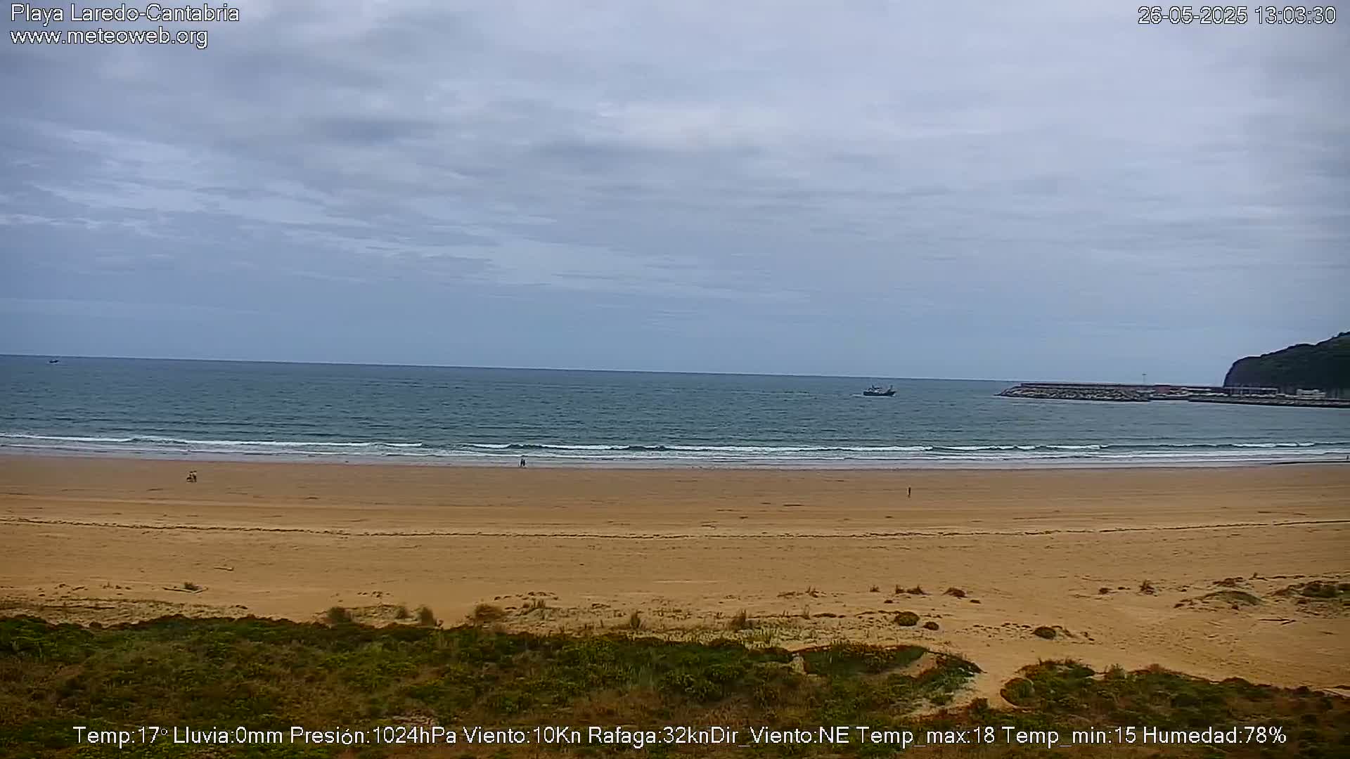 A sandy beach stretches to a calm ocean under an overcast sky, with a few people visible on the sand and a distant boat on the water.