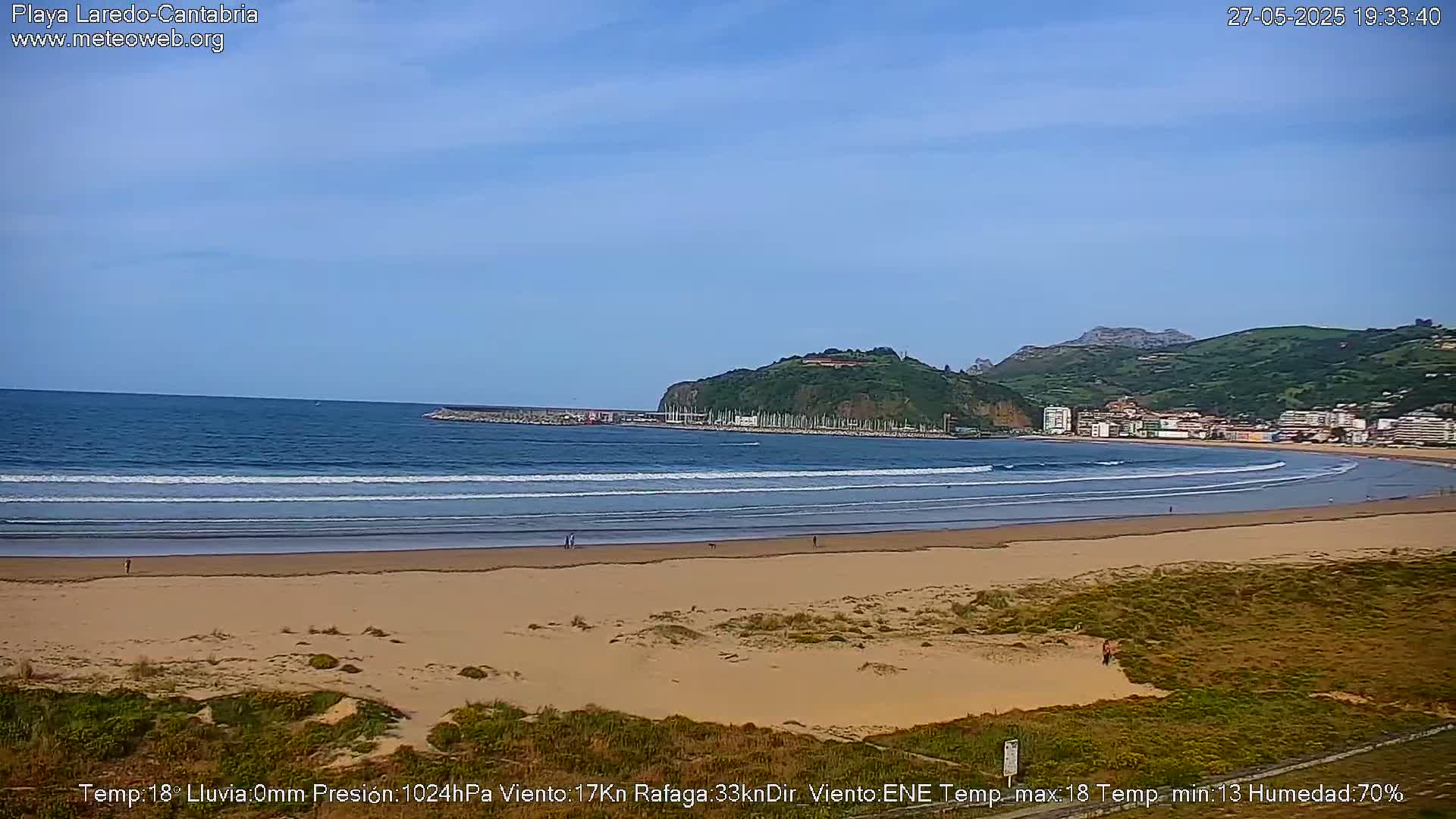 A sandy beach with gentle waves laps the shore under a clear blue sky, backed by a town nestled against a verdant hillside.