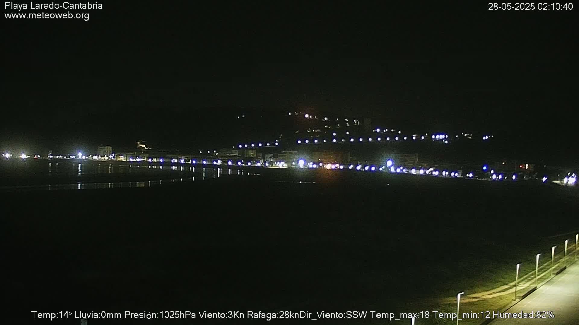 A nighttime view of a dark beach with lights from buildings and vehicles visible in the distance.