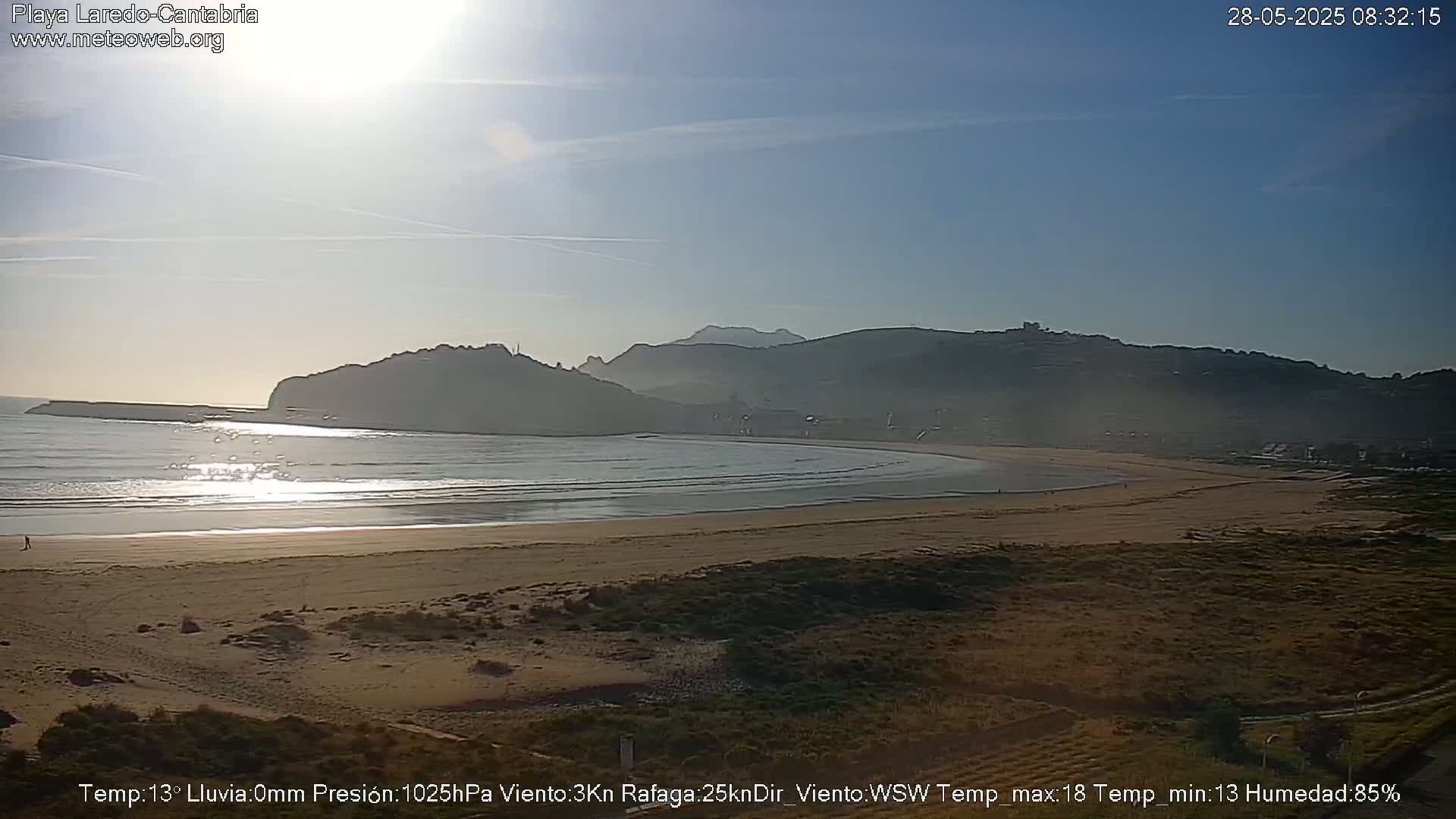 A sandy beach with calm waves stretches beneath a hazy, sunny sky, backed by low, dark hills.