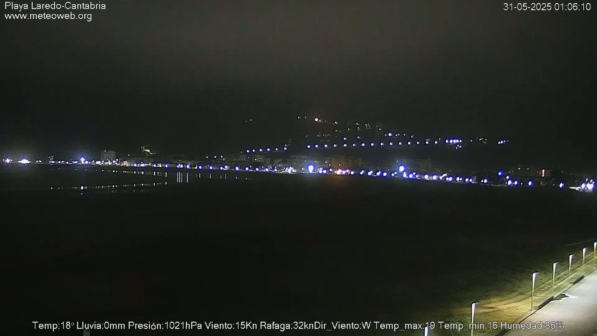 A nighttime view of a coastal town with lights reflecting on calm water under a dark, clear sky.
