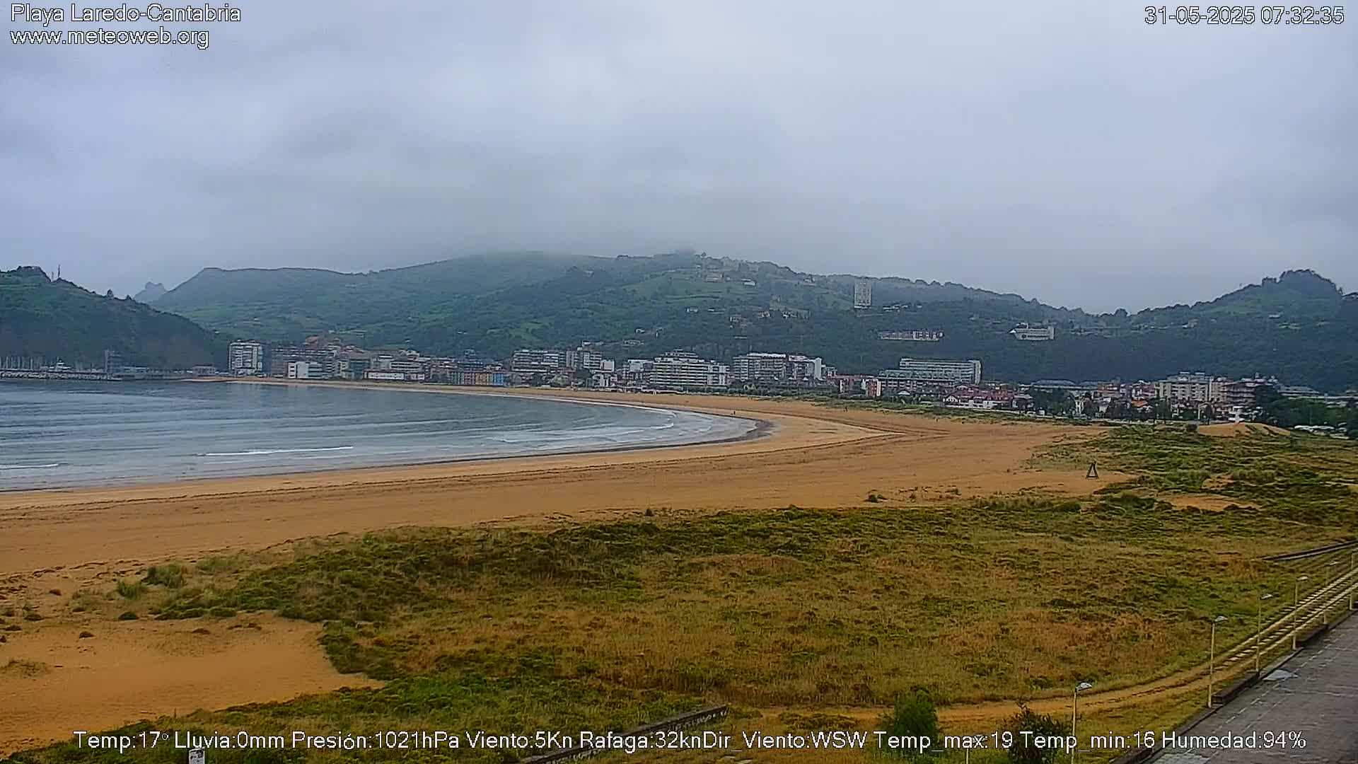 A sandy beach curves gently along a calm bay, backed by a town nestled against a range of low, green hills under an overcast sky.