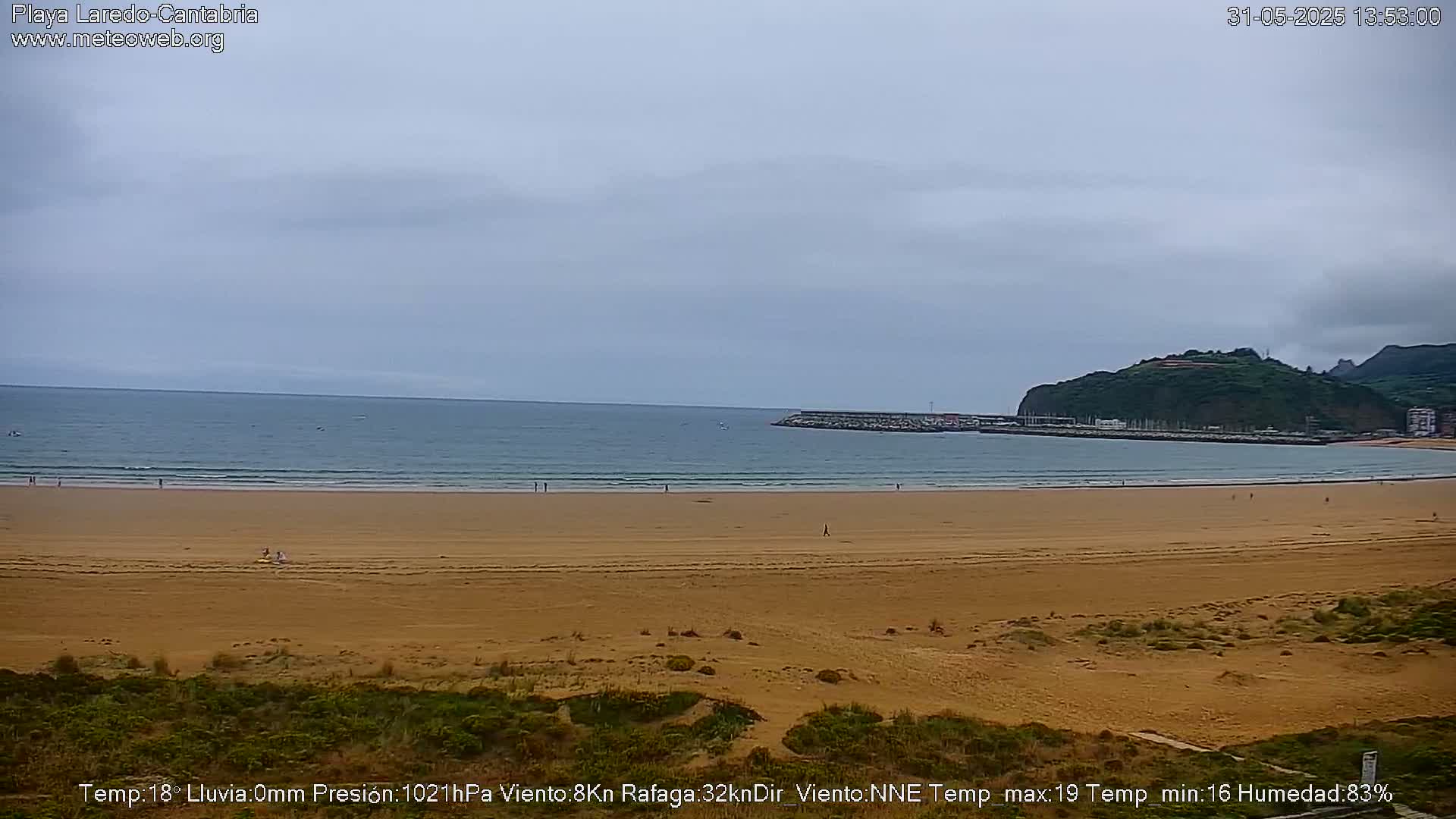A wide sandy beach stretches to a calm sea under an overcast sky, with a few people scattered along the shore and a harbor visible in the distance.
