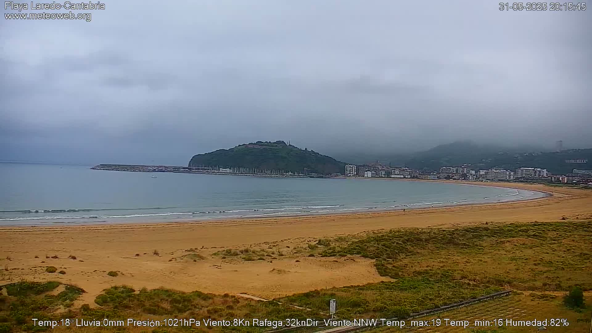 A sandy beach curves around a calm bay under a cloudy sky, with a green hill and town visible in the distance.