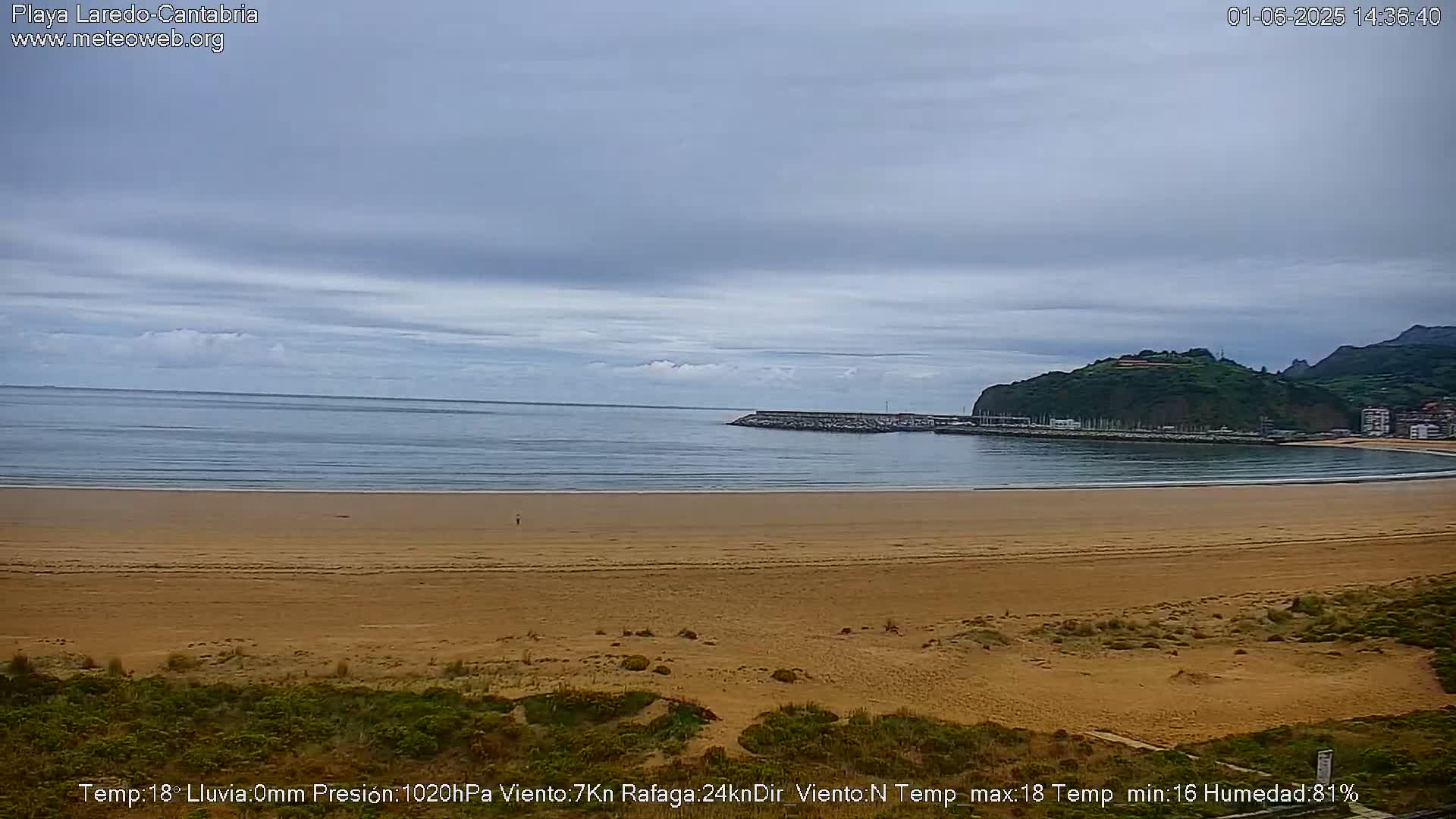 A sandy beach stretches to a calm ocean under an overcast sky, with a small harbor and a hill visible in the distance.